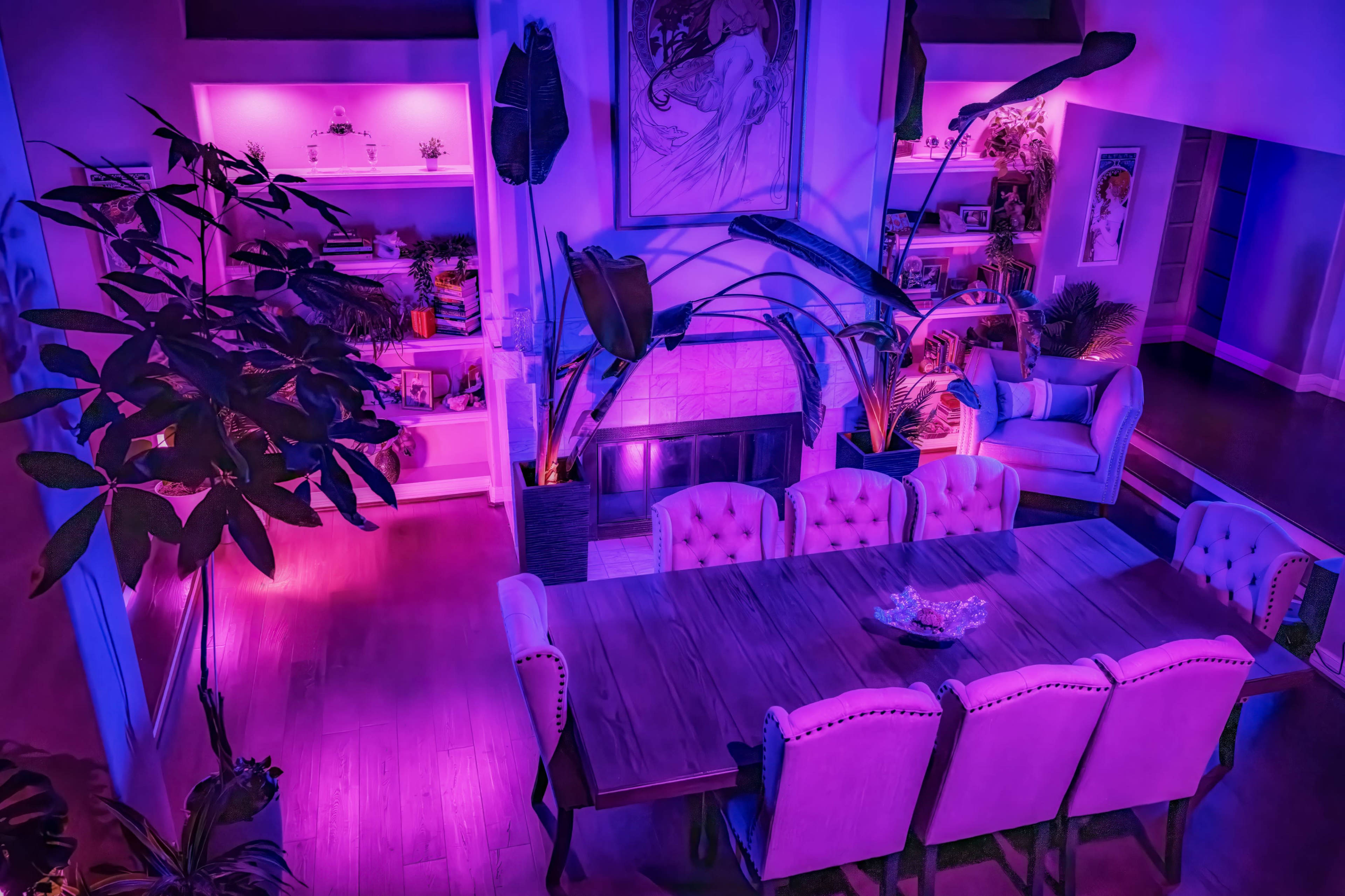 A dining room features a long wooden table surrounded by upholstered chairs, illuminated by purple lighting, while a cozy sitting area is visible in the background.