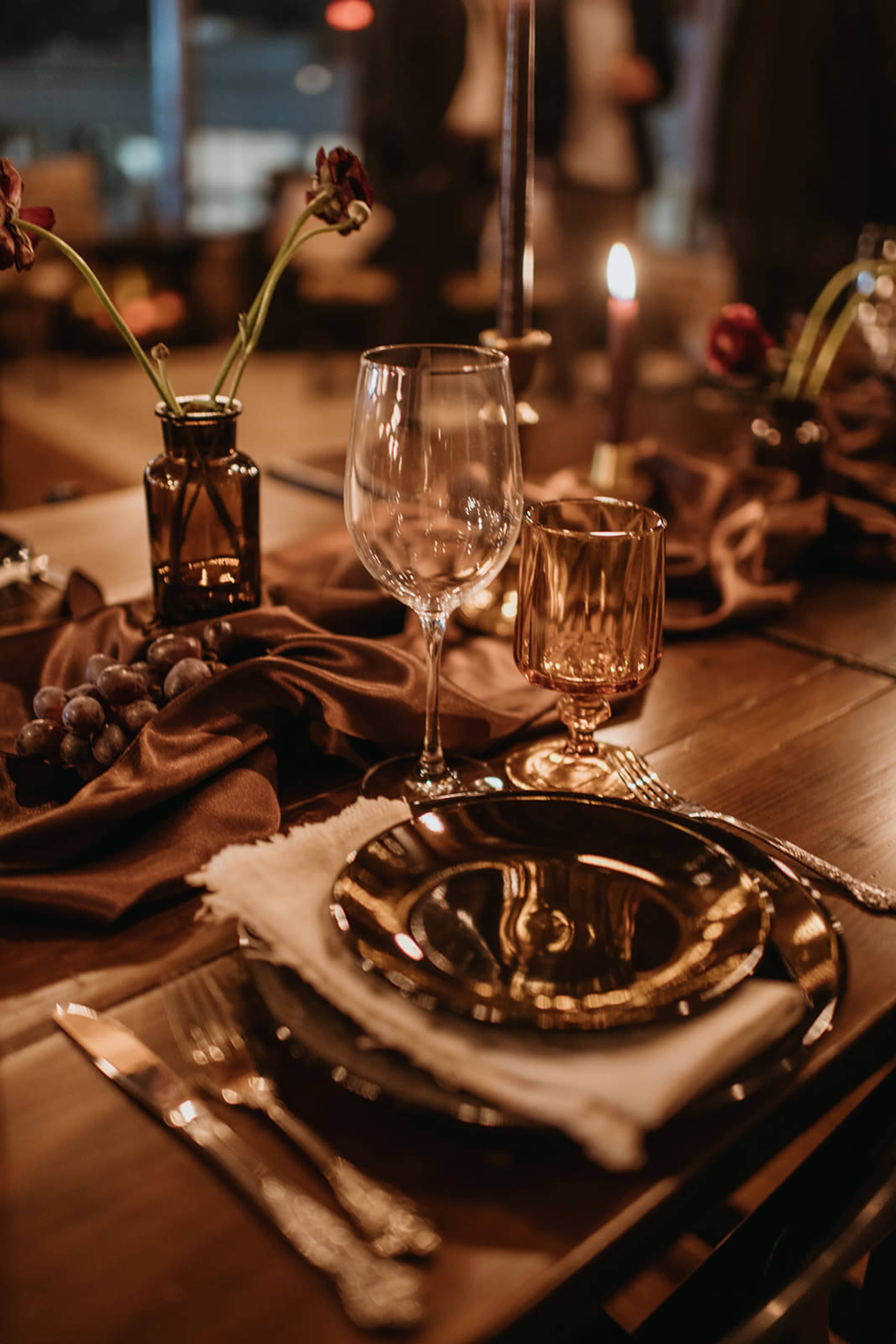 The image shows a beautifully arranged dining table featuring elegant glassware, dark plates, and decorative flowers, all set against a backdrop of soft candlelight.