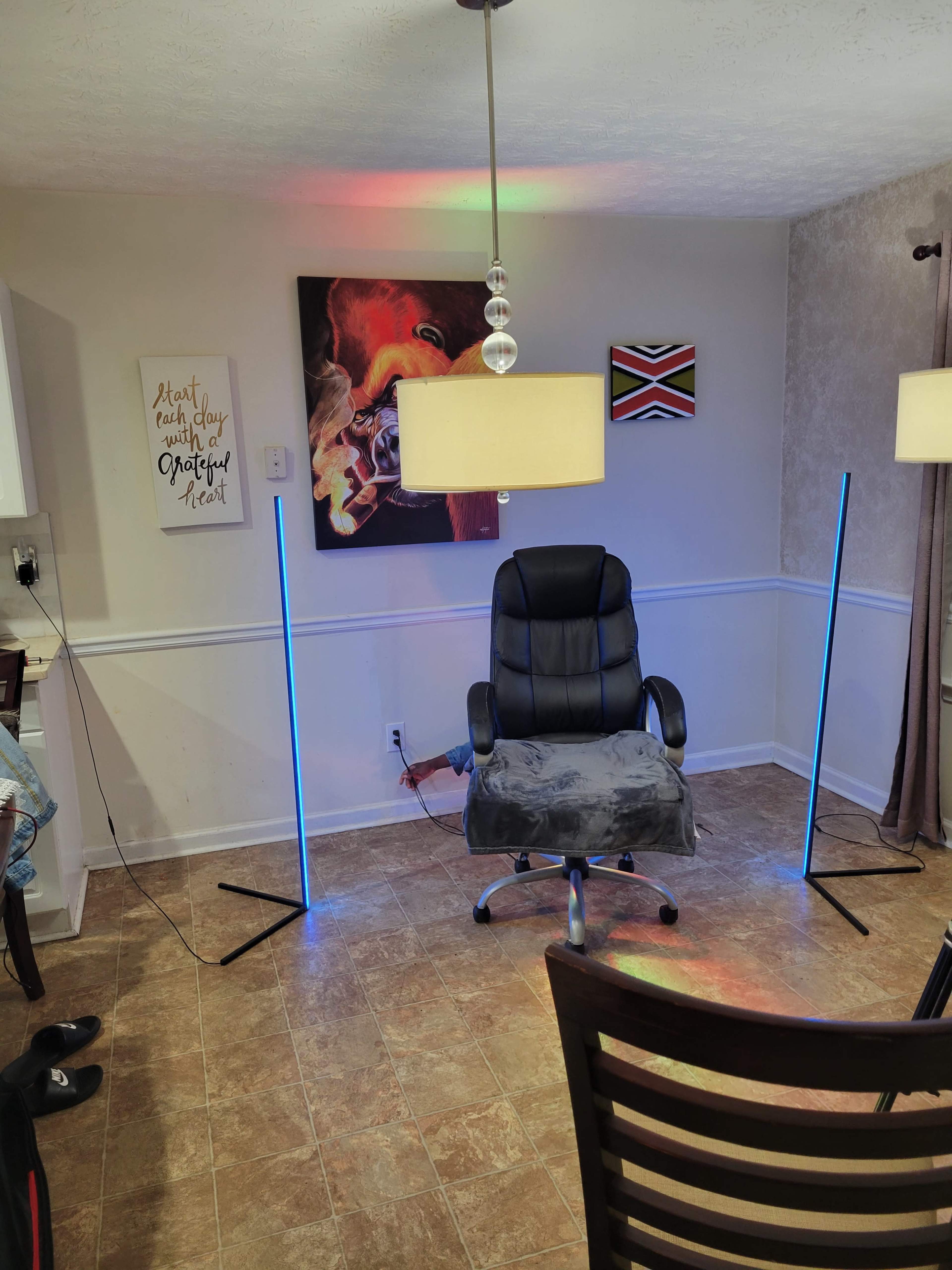 The image shows a small room with a black rolling chair in the center, flanked by two vertical light stands emitting blue light, and adorned with framed artworks on the walls.