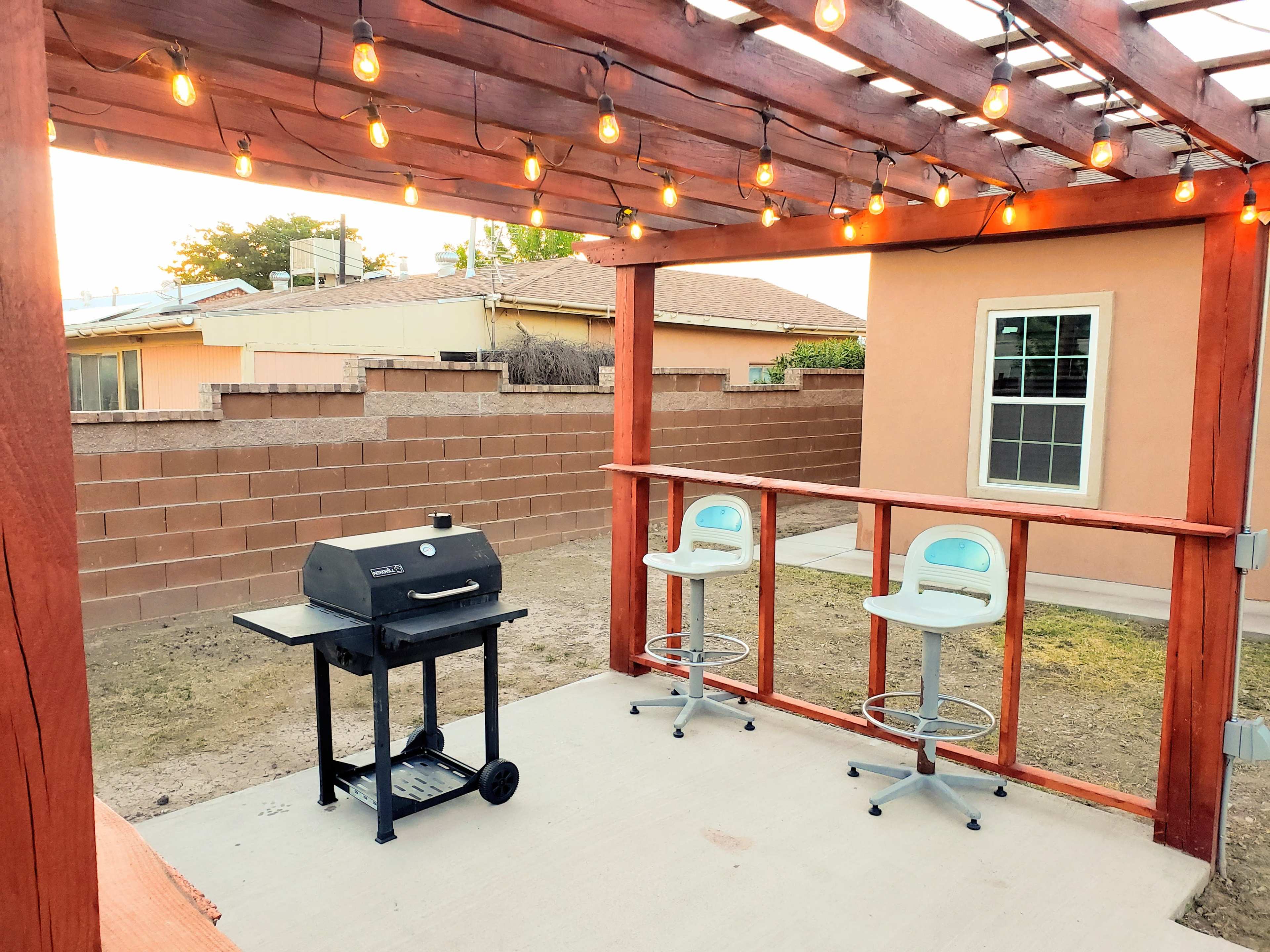 A backyard patio area with a charcoal grill, two white bar stools, and string lights overhead.