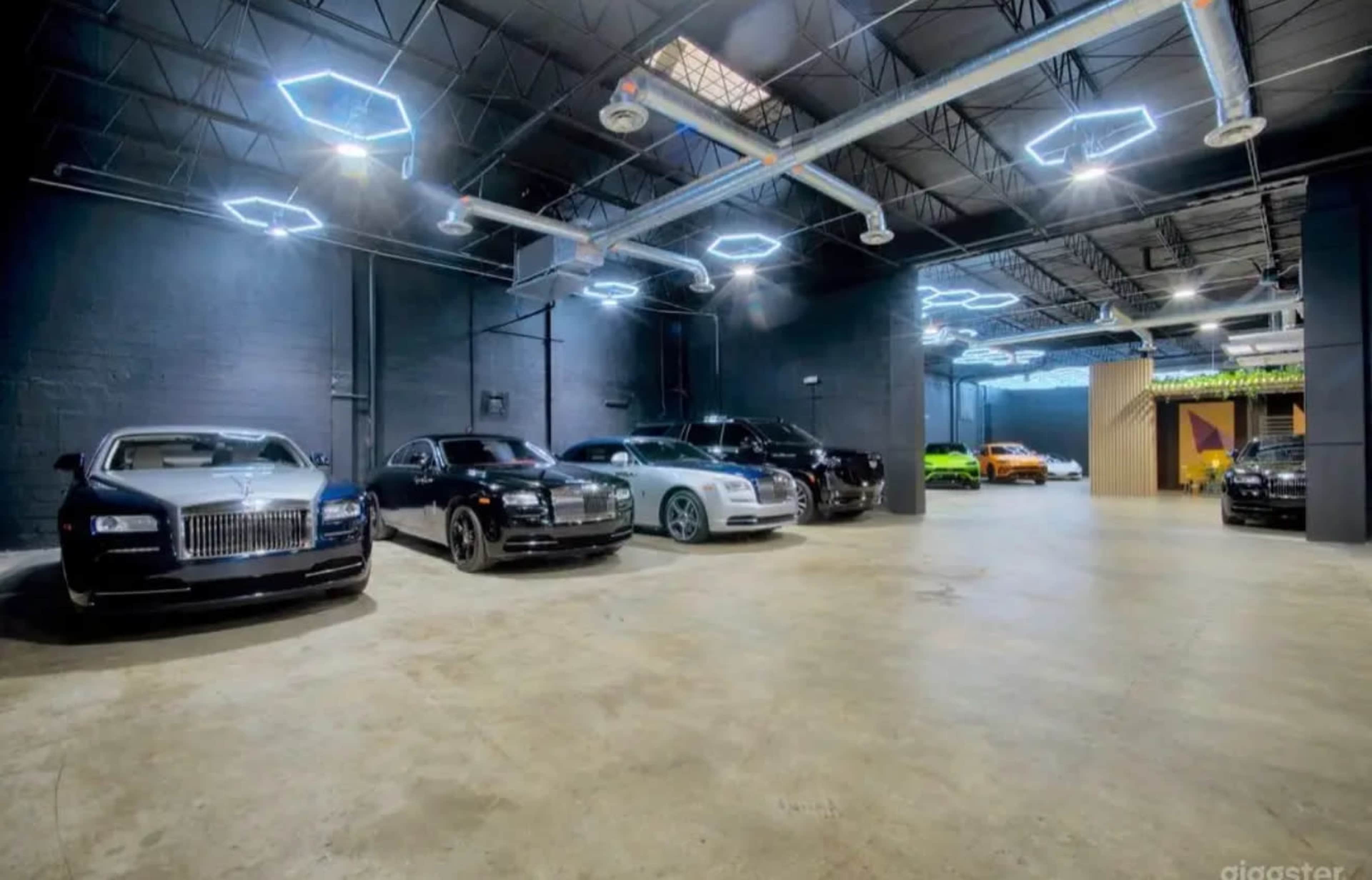 The image shows a spacious garage with several luxury cars lined up on one side, illuminated by modern ceiling lights.