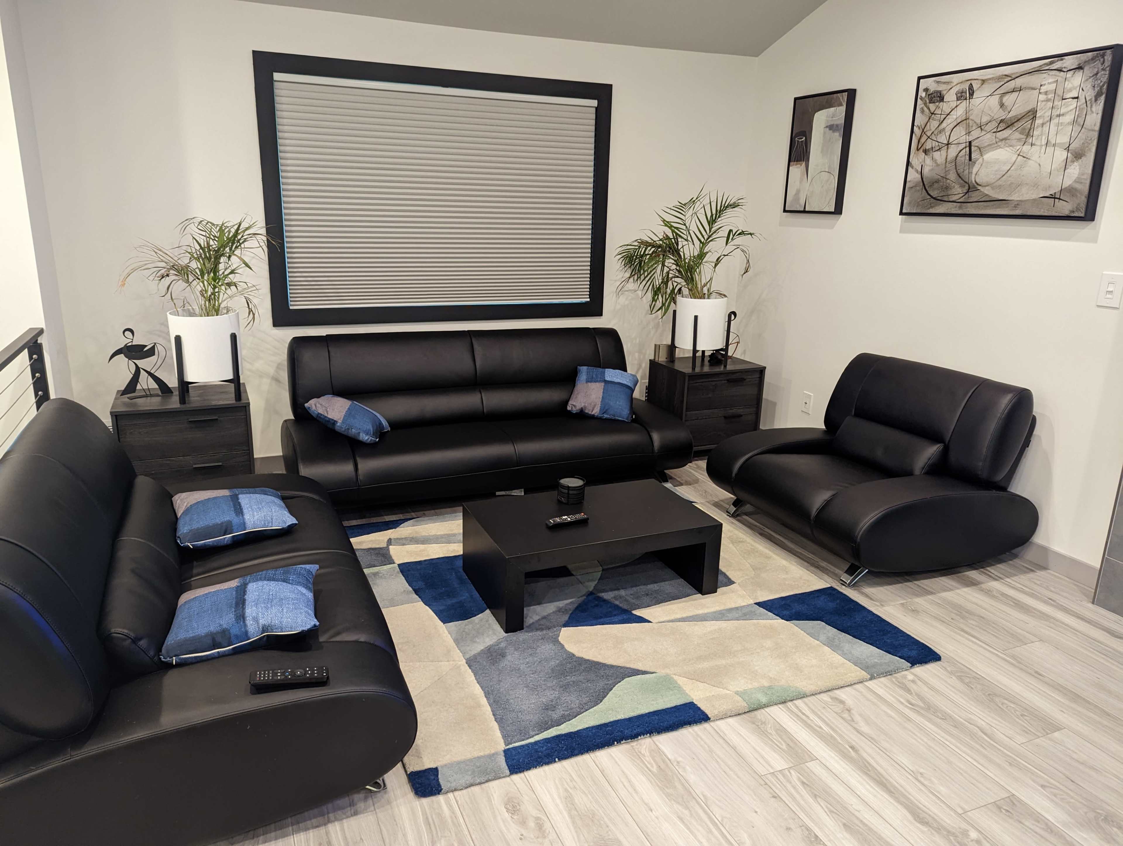 A modern living room features black leather furniture, a coffee table, and a patterned area rug, surrounded by plants and wall art.