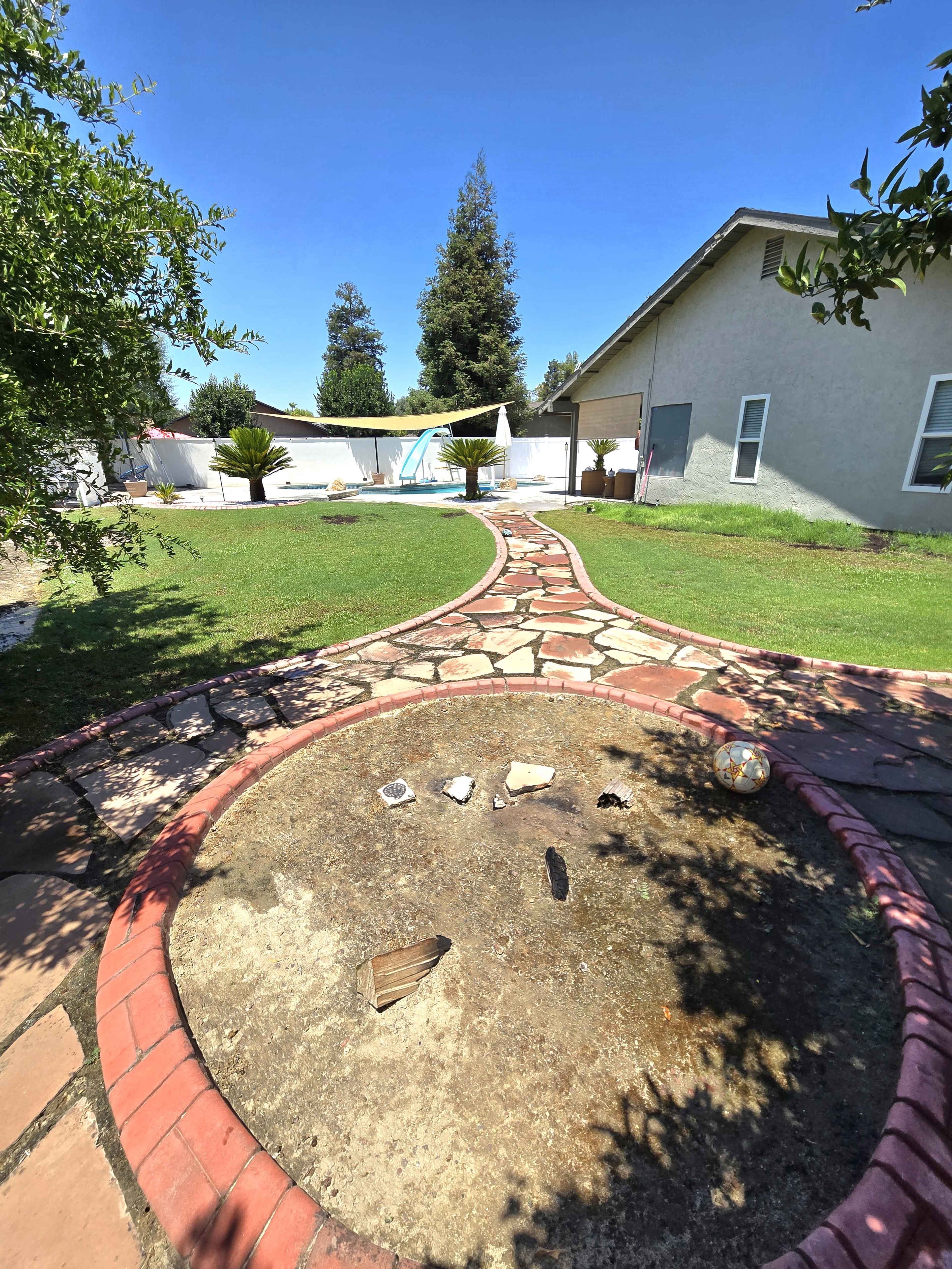 A stone pathway curves through a yard leading to a pool area surrounded by greenery and a house in the background.