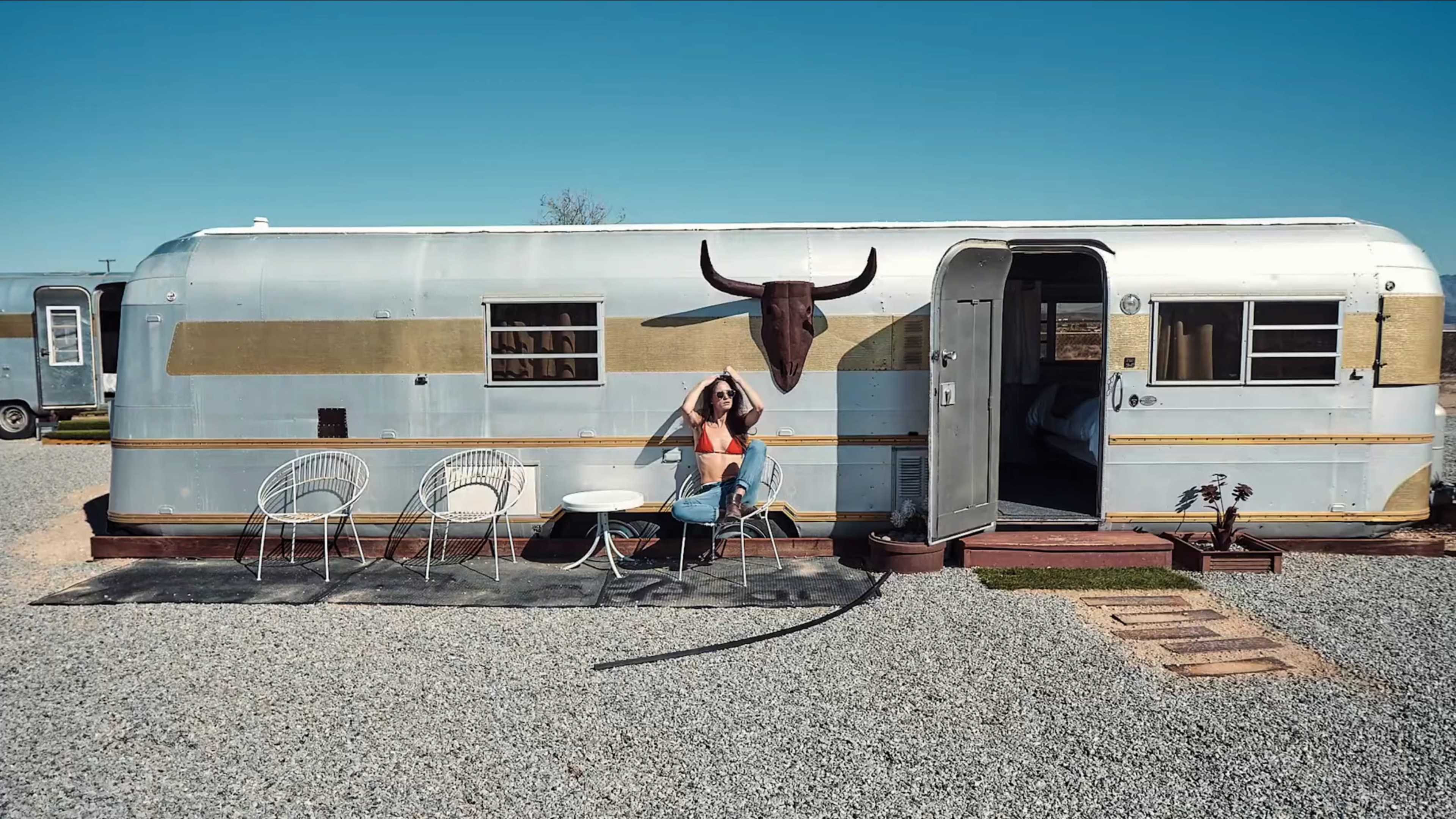A woman sits at a table in front of a vintage silver trailer adorned with a bull's skull, surrounded by gravel and outdoor furniture.