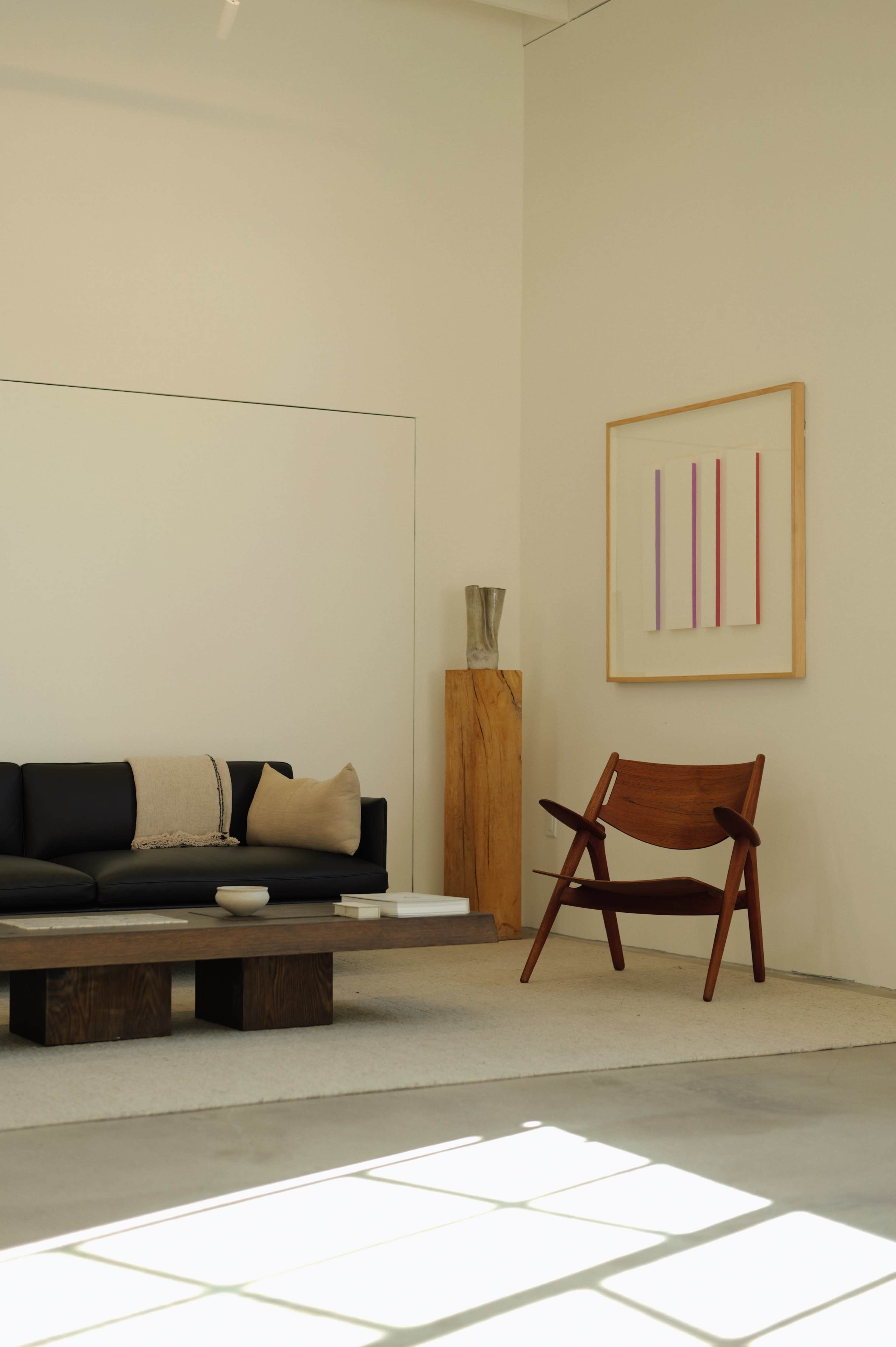 The image features a minimalist interior with a black sofa, a wooden coffee table, a wooden sculpture, a framed artwork on the wall, and a brown accent chair.