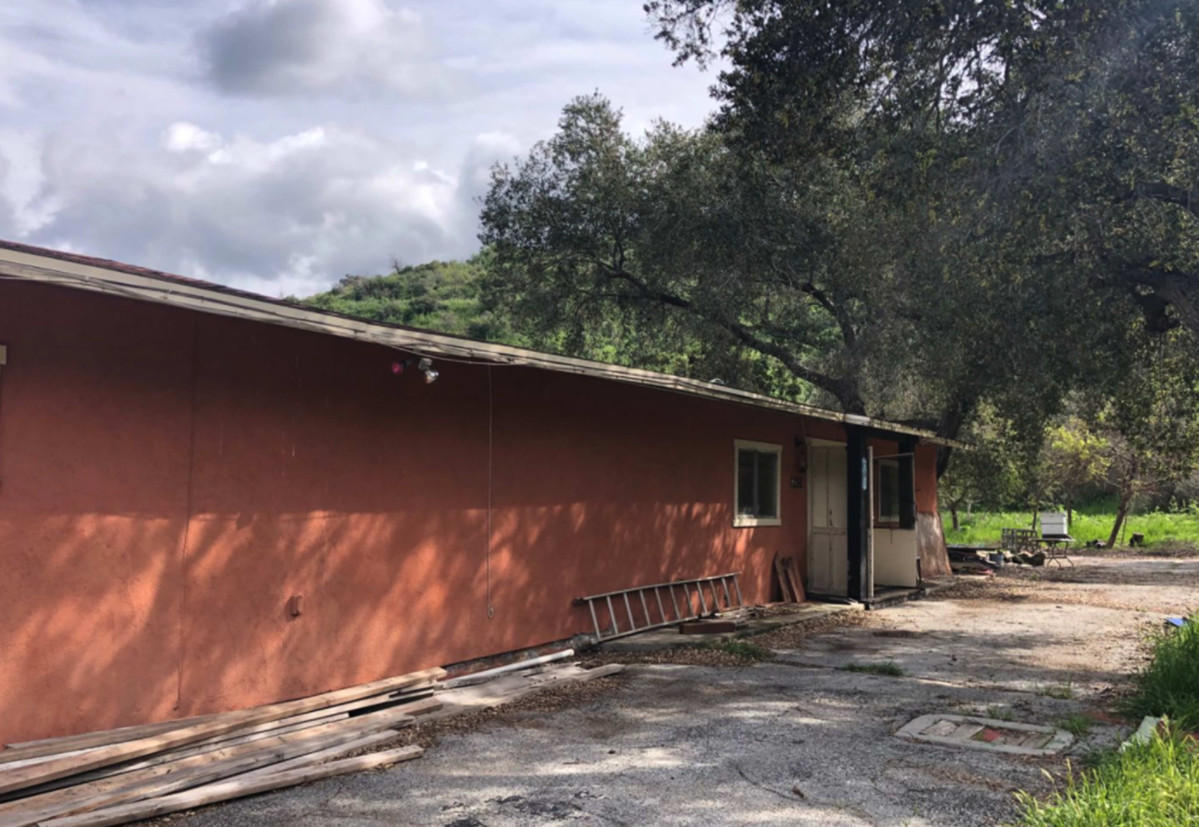 A single-story, orange-brown building stands alongside a gravel path, surrounded by trees and green hills in the background.