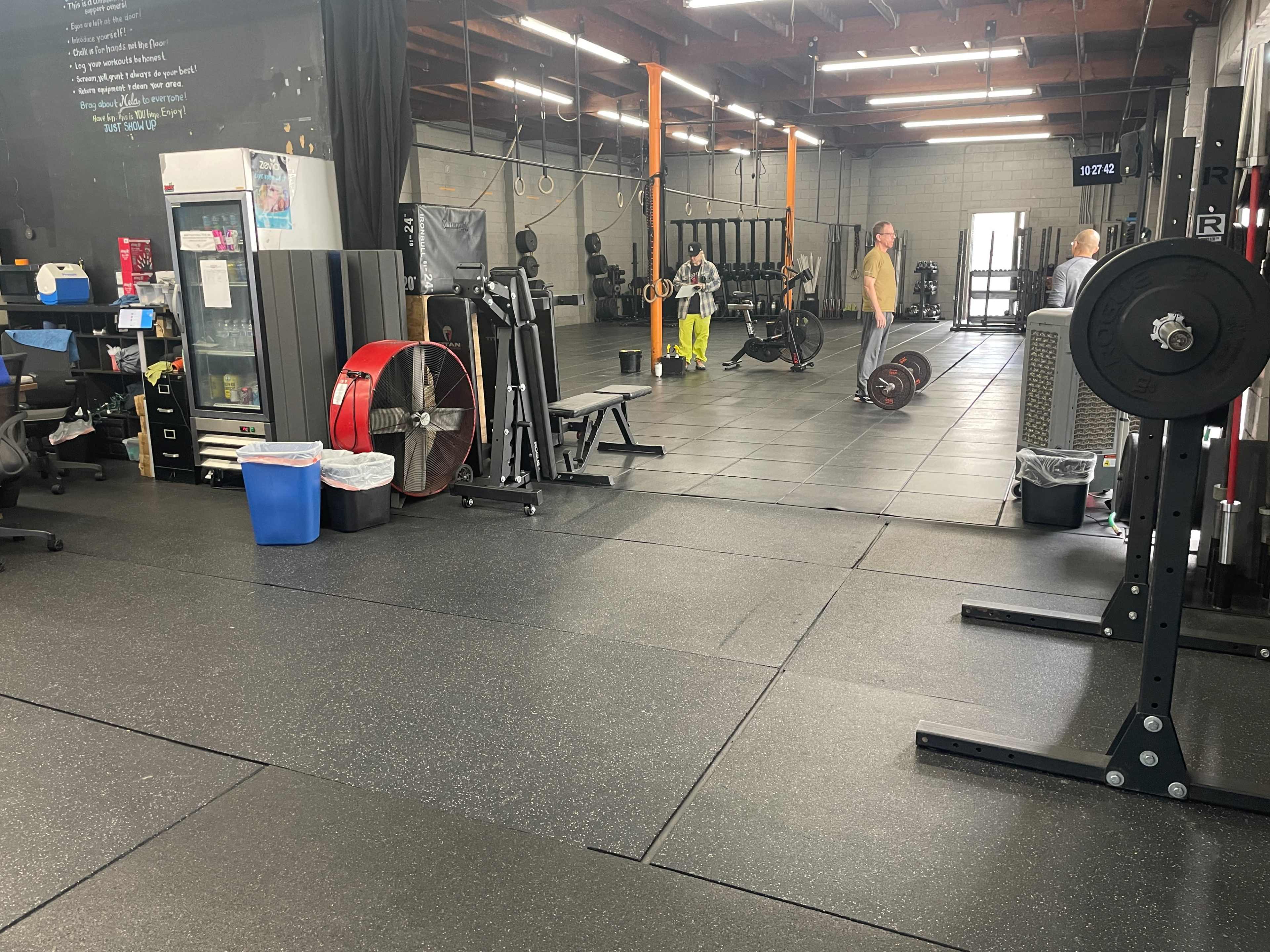 A gym interior with exercise equipment, including free weights, mats, and a fan, alongside two people engaging in an activity in the background.