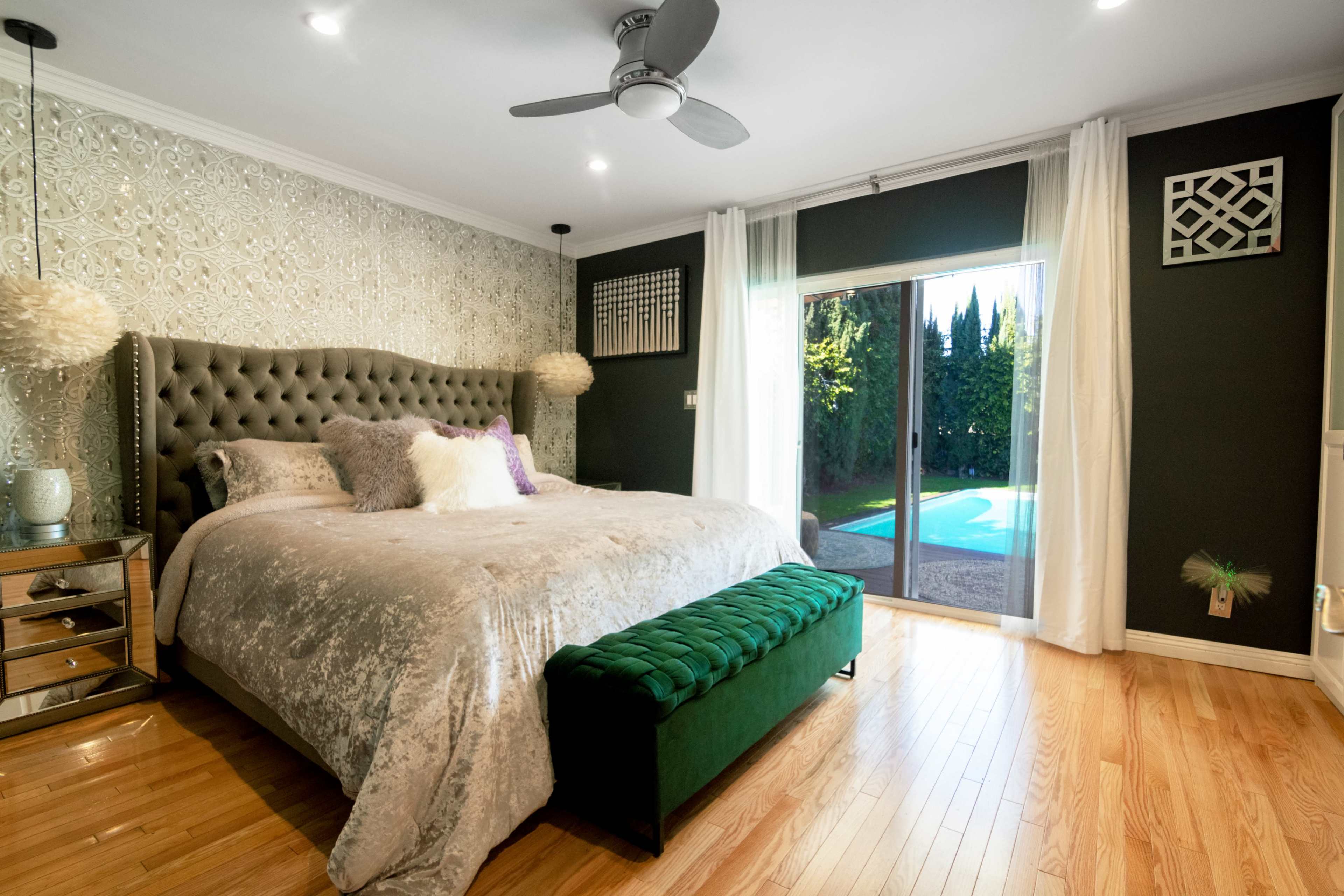 The image shows a modern bedroom with a large upholstered bed, decorative pillows, a green velvet bench, and a sliding glass door that opens to a pool area outside.