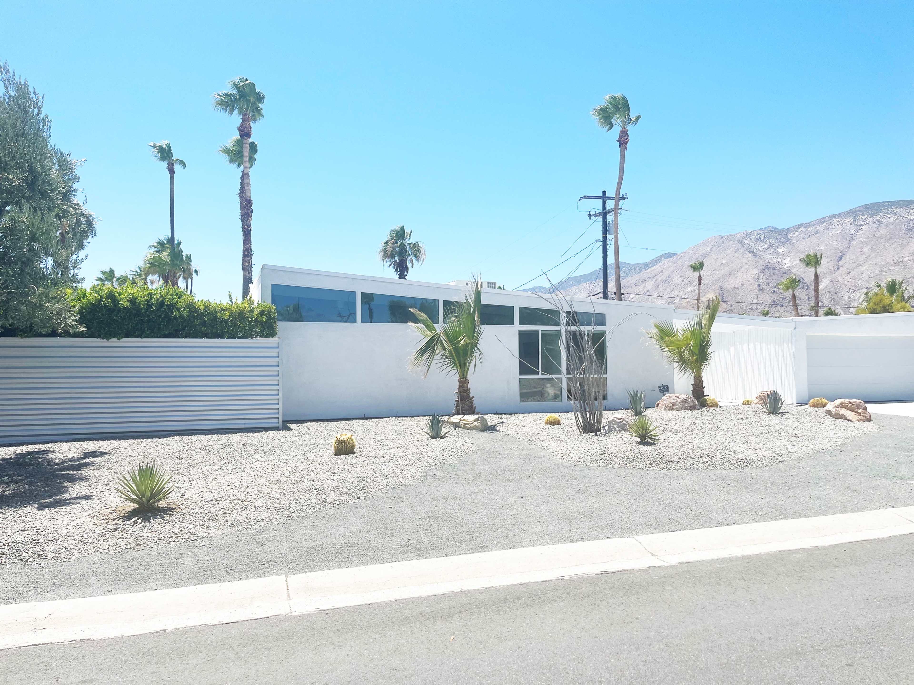 The image shows a modern, one-story house surrounded by palm trees and gravel landscaping in a desert setting.