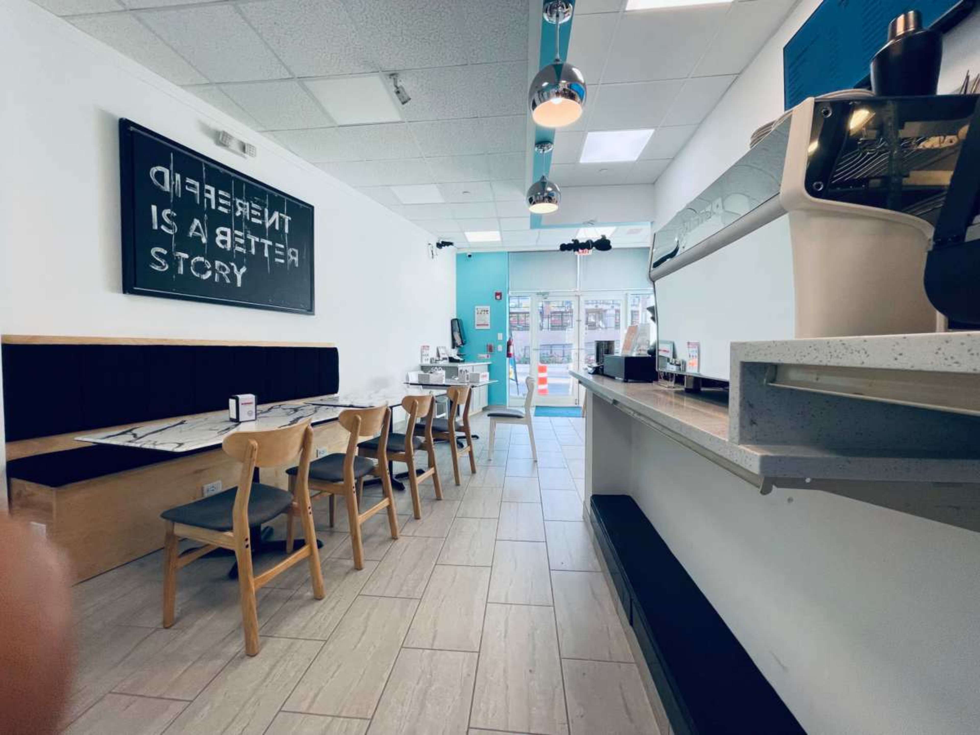 The image shows the interior of a modern cafe with a row of chairs and tables on one side and a coffee counter on the other.