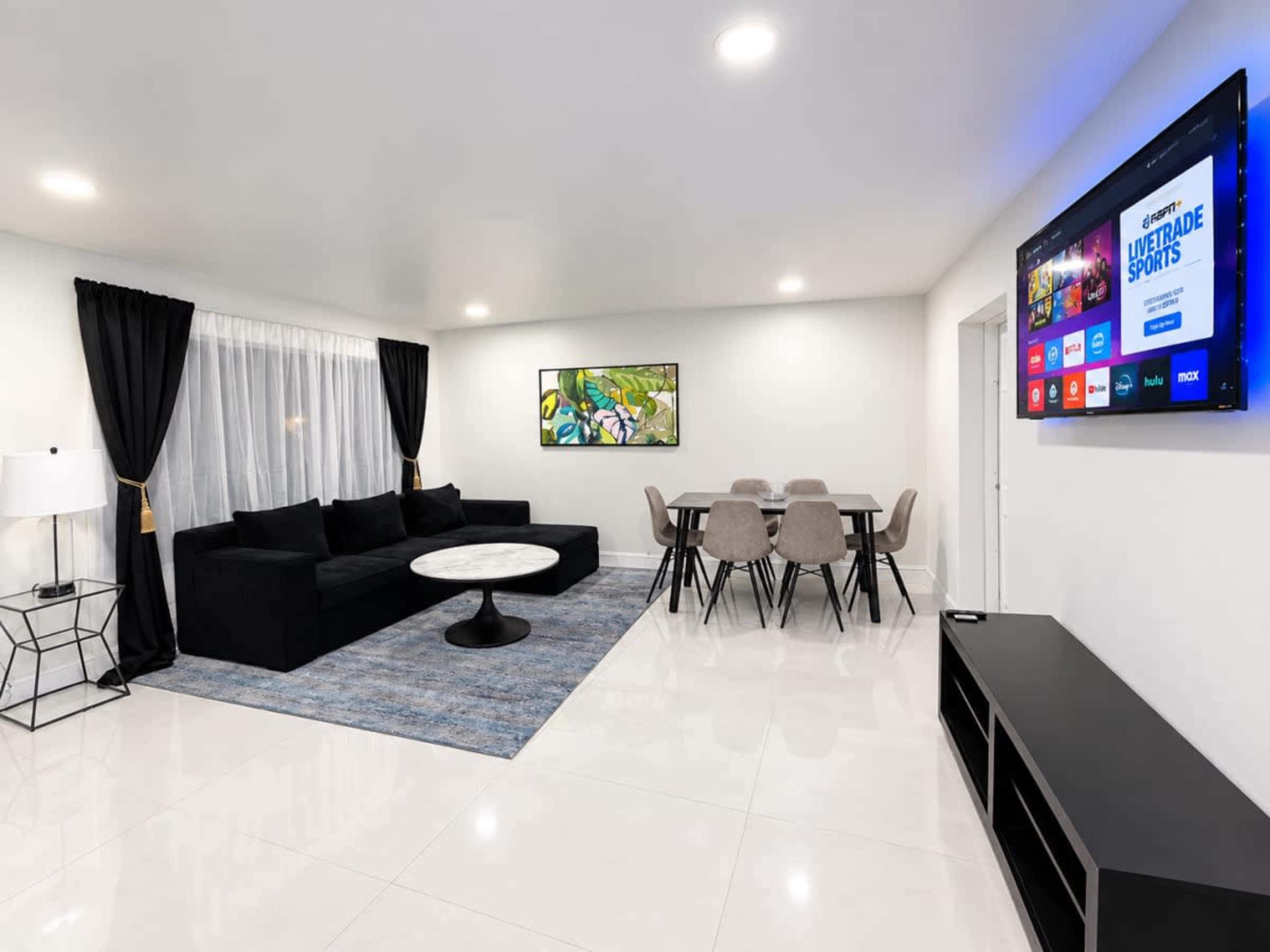 The image shows a modern living room with a black sectional sofa, a round marble coffee table, a dining table with gray chairs, and a wall-mounted television.