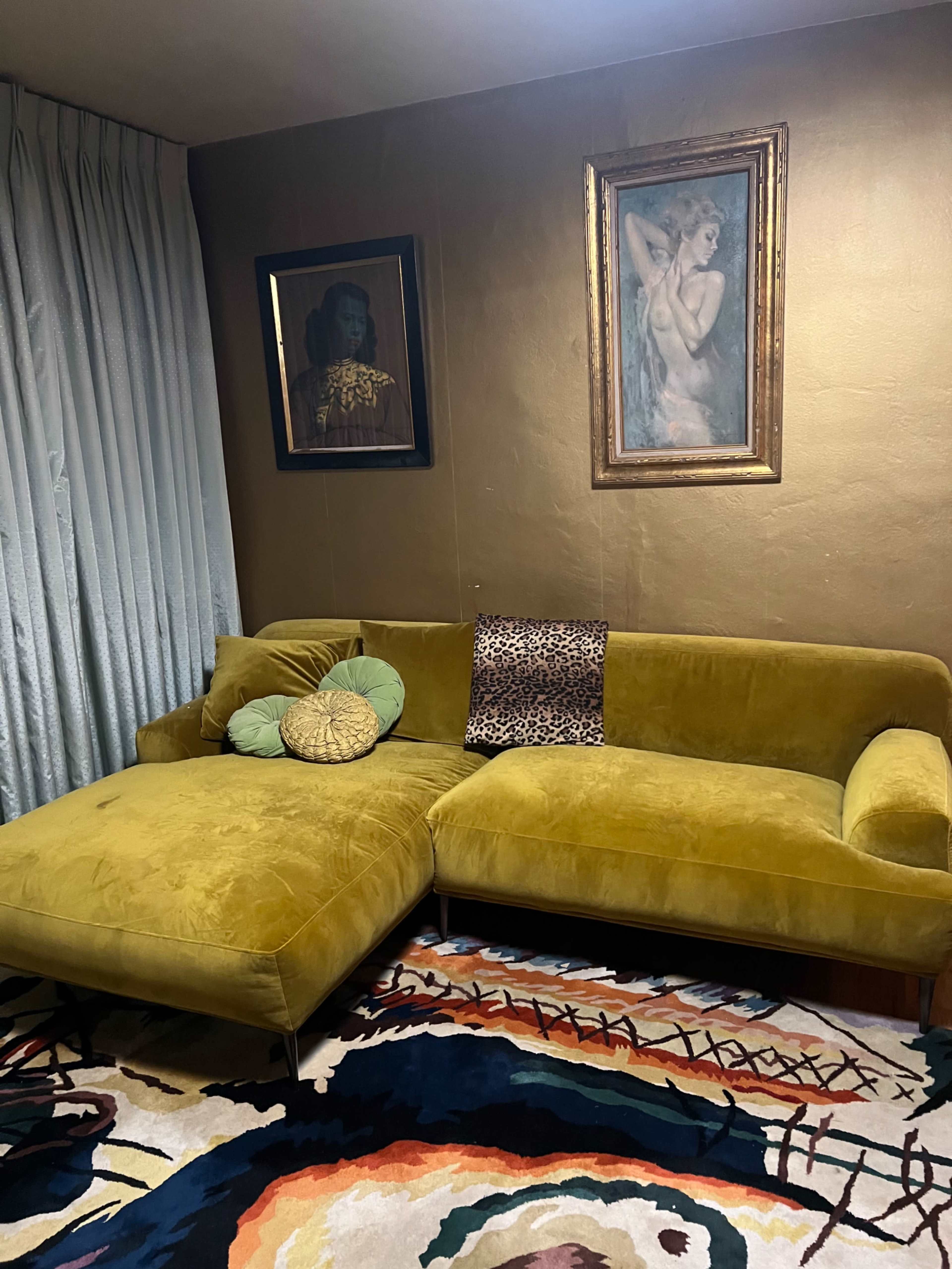 The scene features a green sectional sofa accompanied by a decorative pillow, positioned near a gold-painted wall, with two framed artworks above it and a colorful patterned rug underneath.