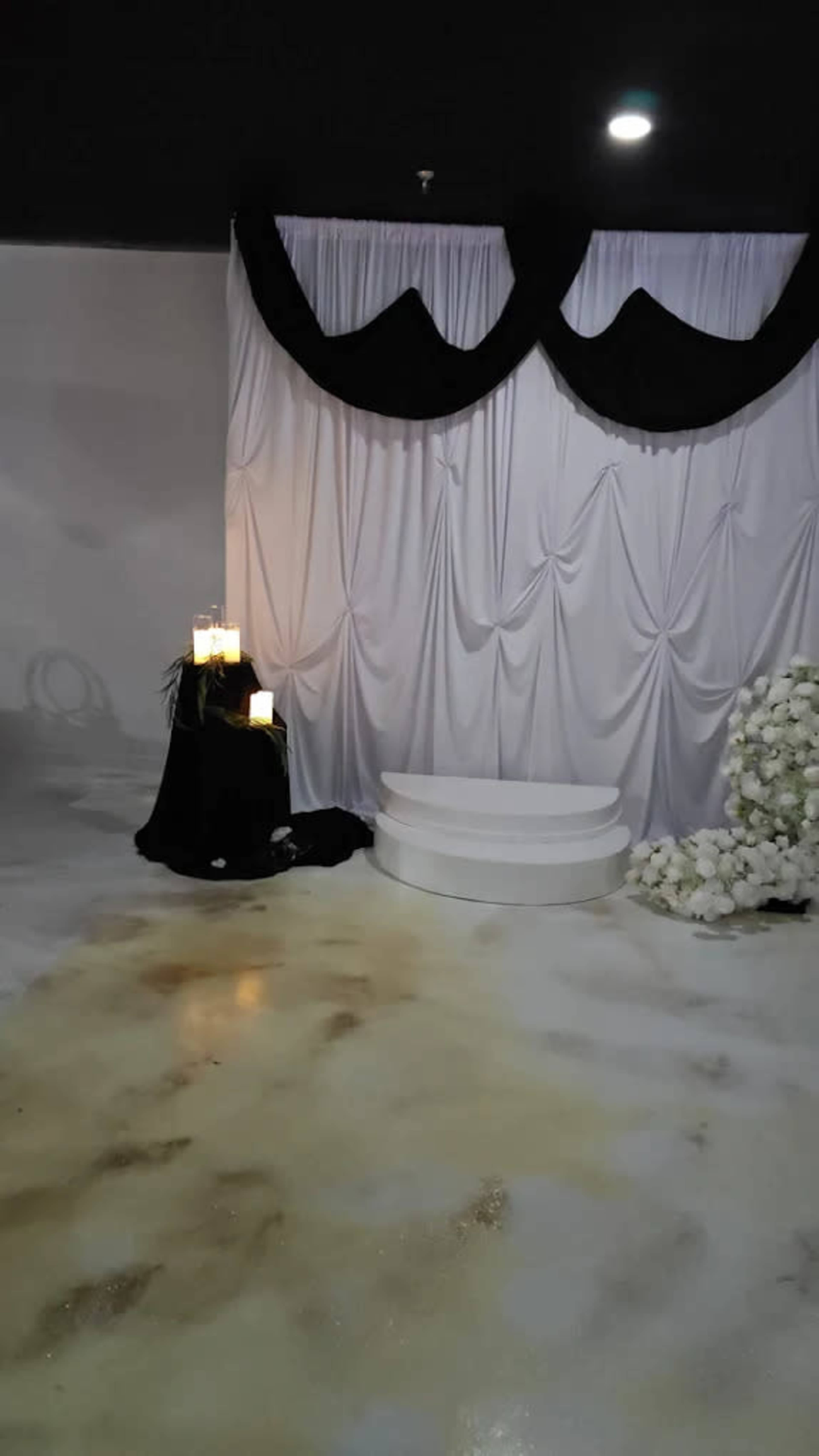 The image shows a decorated stage with white draped fabric, black fabric accents, a set of candles on a pedestal, and a floral arrangement on the side.