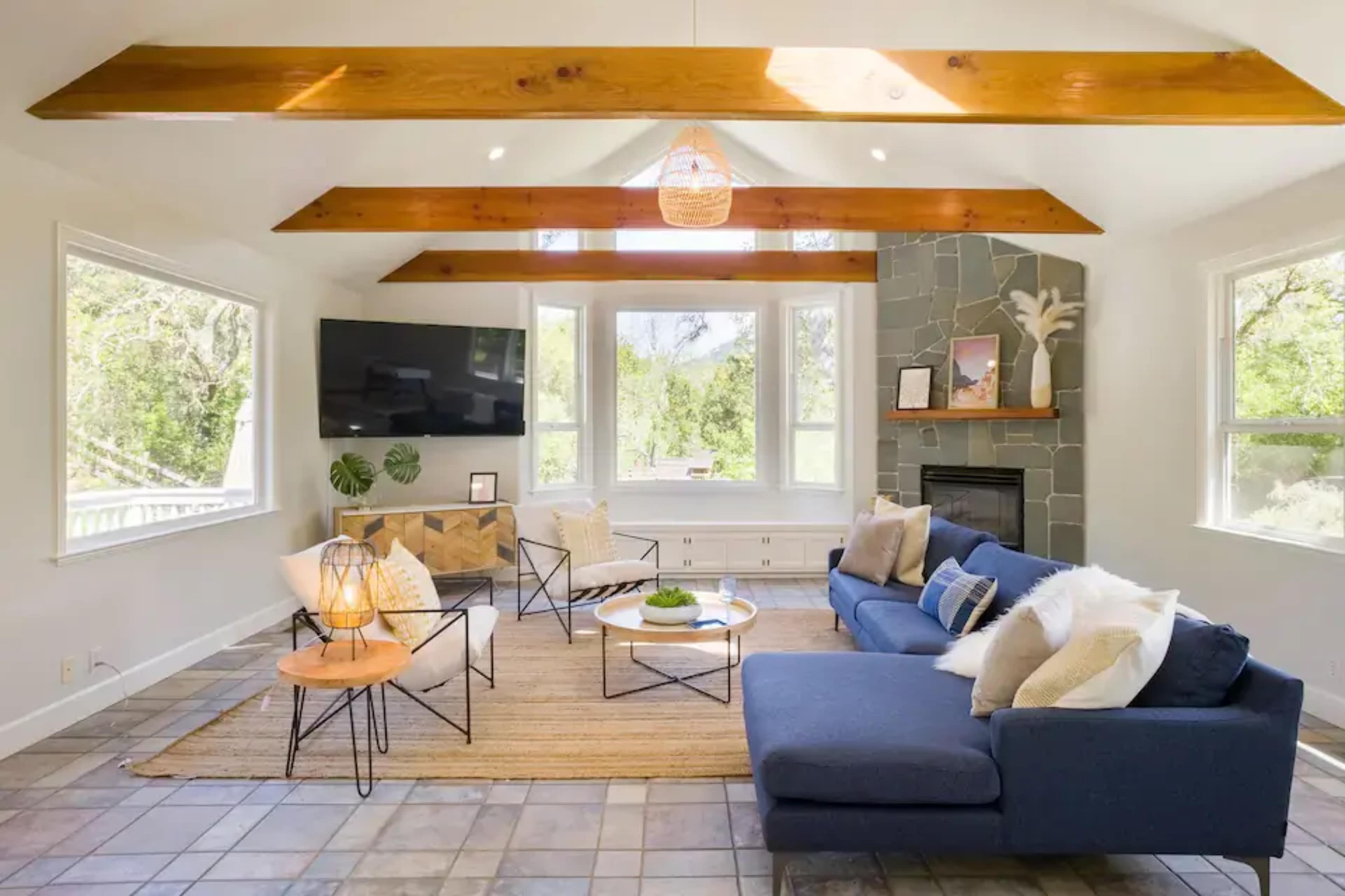 The living room features a blue sofa, a television mounted on the wall, and large windows that provide a view of the outdoors, all under wooden beam ceilings.