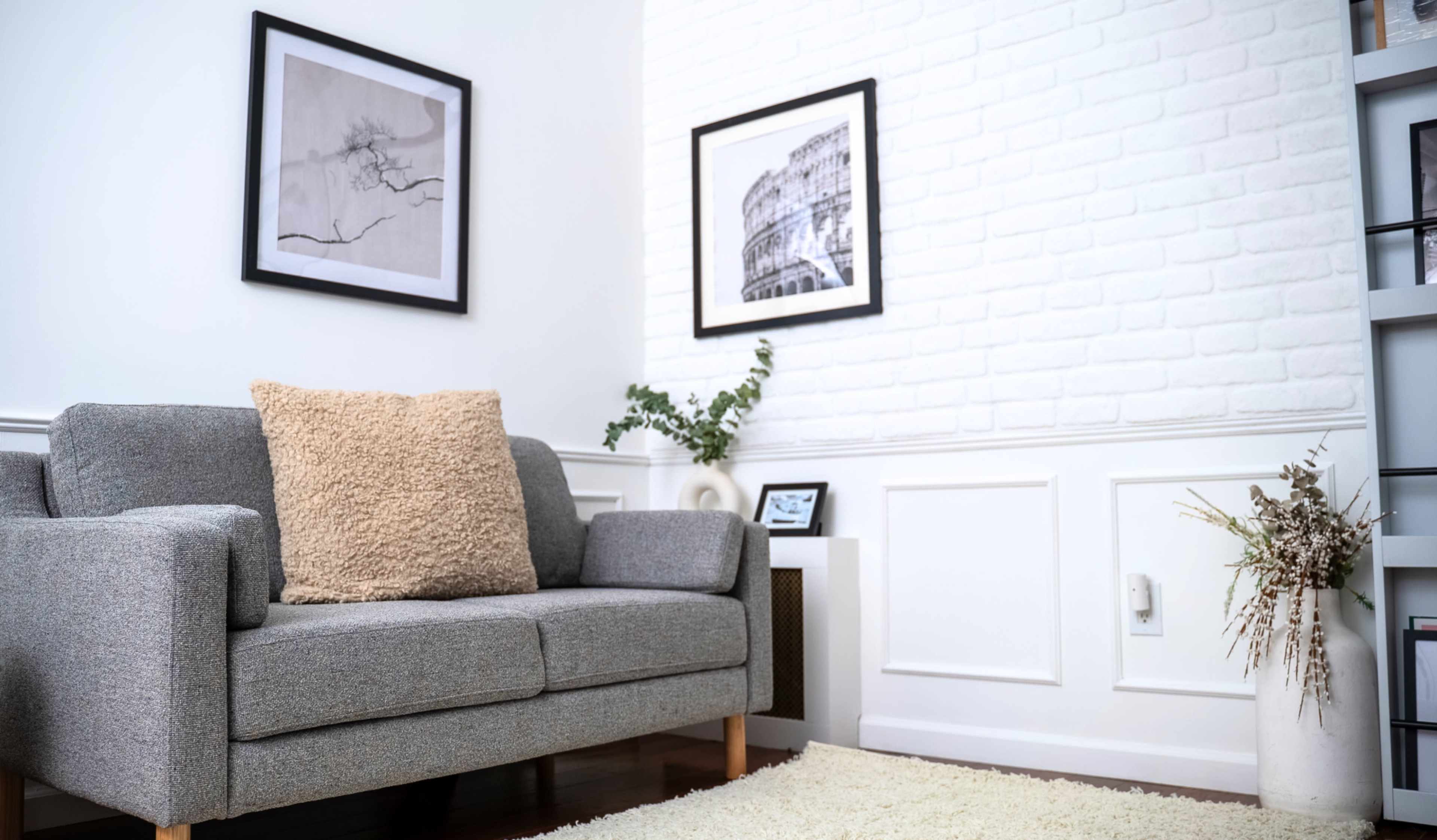 A modern living room features a gray sofa with a textured throw pillow, framed artwork on the walls, and a small potted plant on a side table.