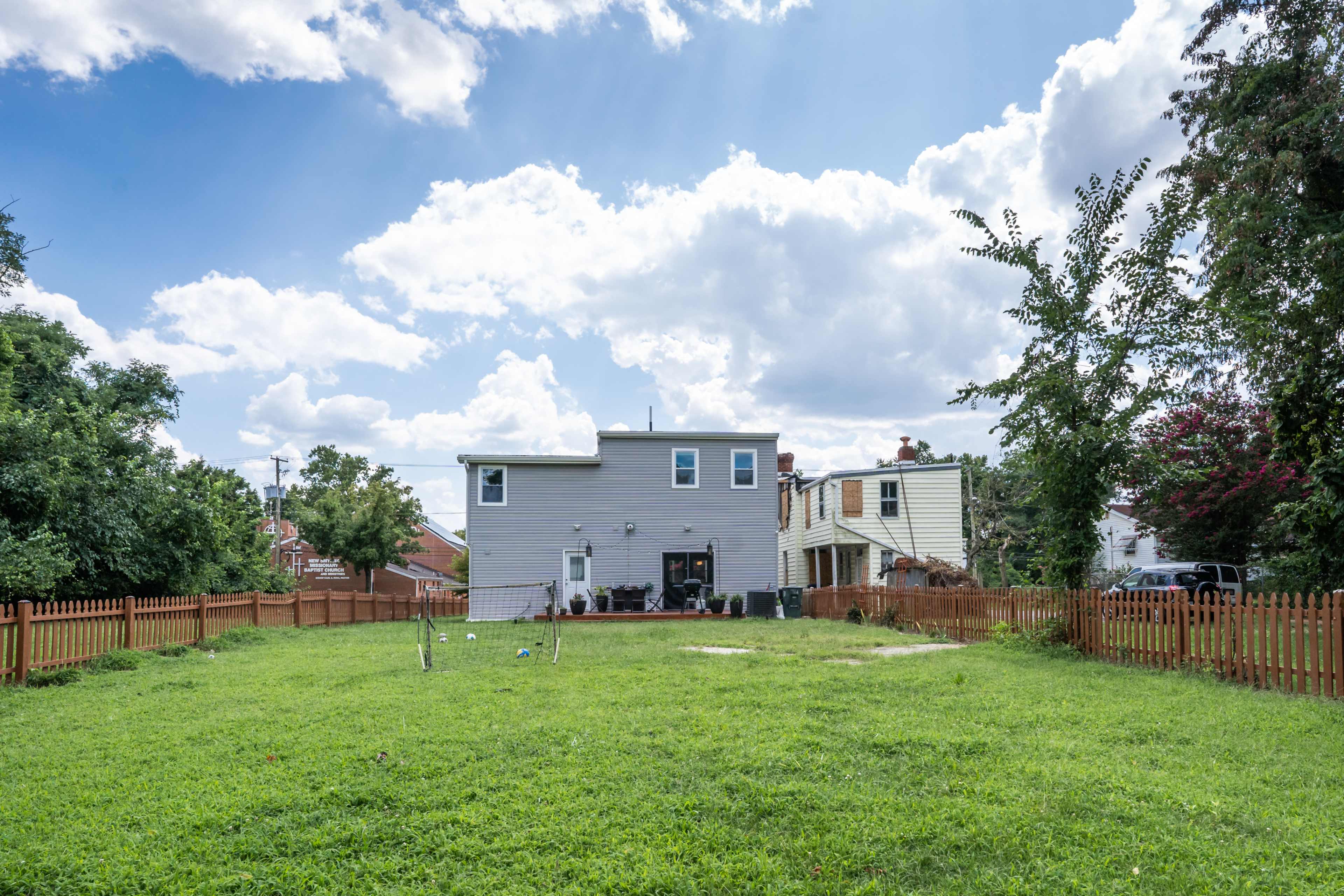 A backyard with a fenced area, featuring a two-story gray house and a grassy lawn under a cloudy sky.