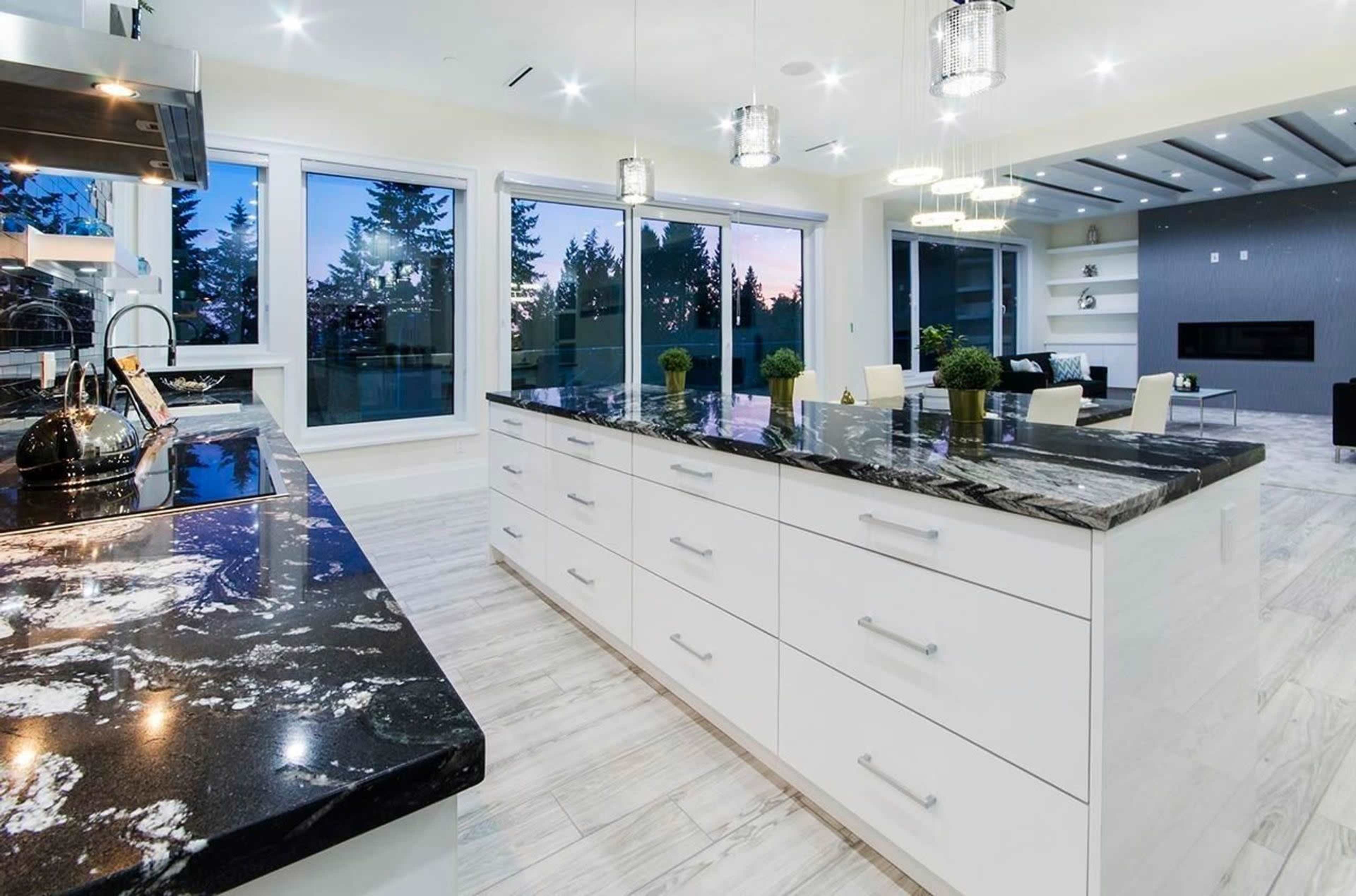 A modern kitchen features a large island with a dark marble countertop and white cabinetry, overlooking a view of trees and a sunset through large windows.