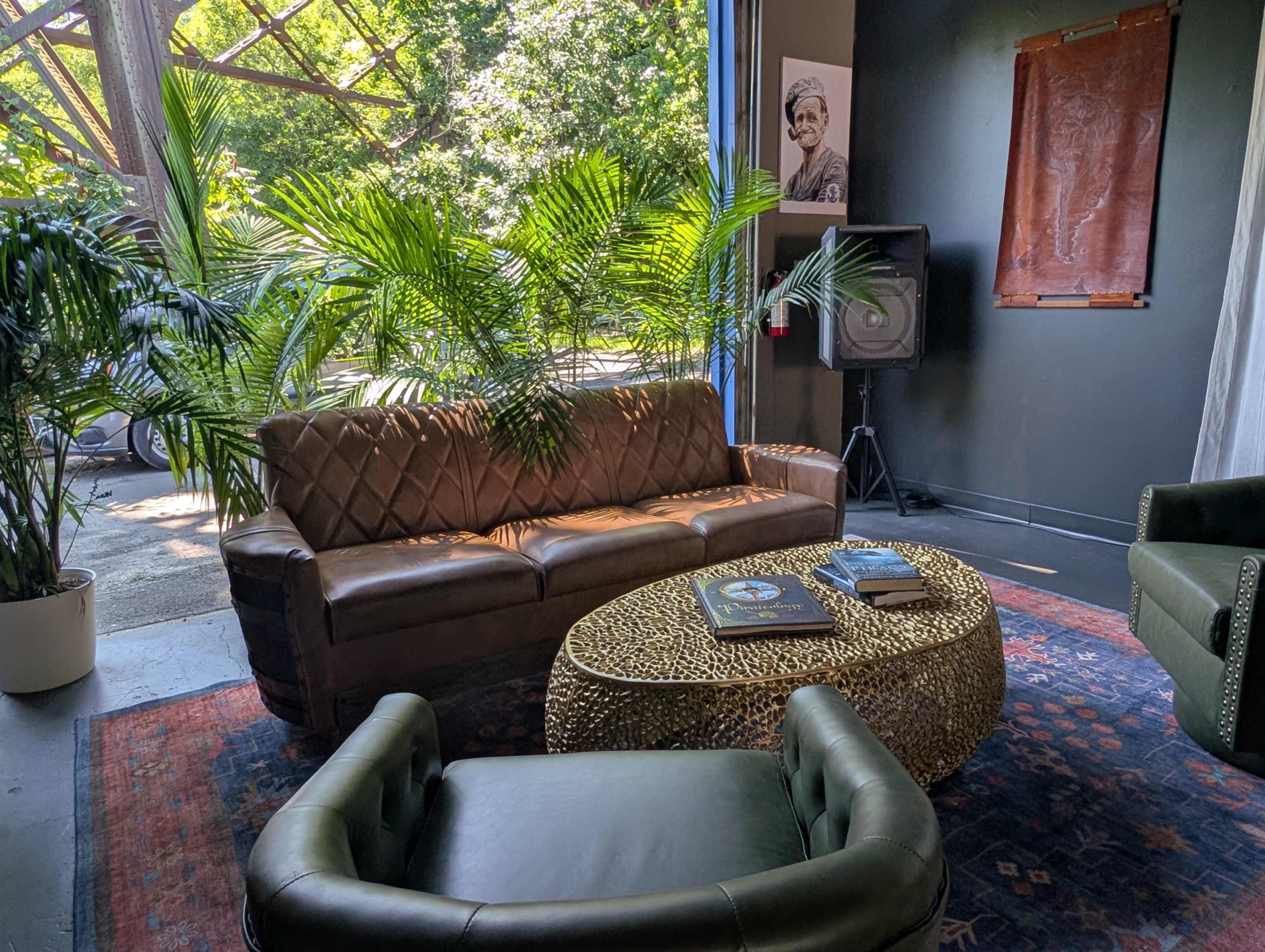 The image shows a cozy lounge area featuring a brown leather sofa, a decorative coffee table with a textured surface, and green plants beside an antique rug.