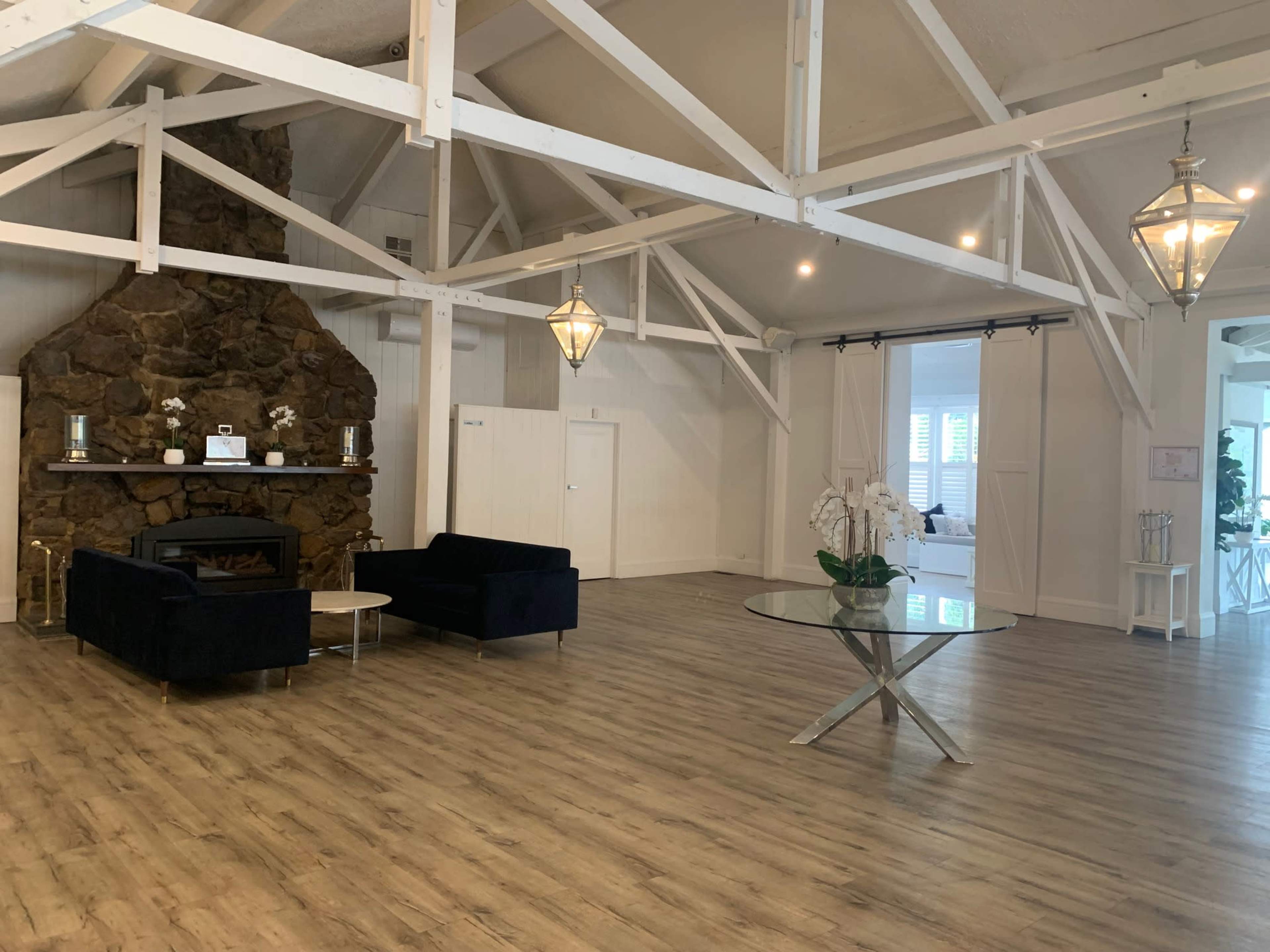 The image shows a spacious interior with a stone fireplace, two dark sofas, a coffee table, and large windows, all under a high ceiling supported by white beams.