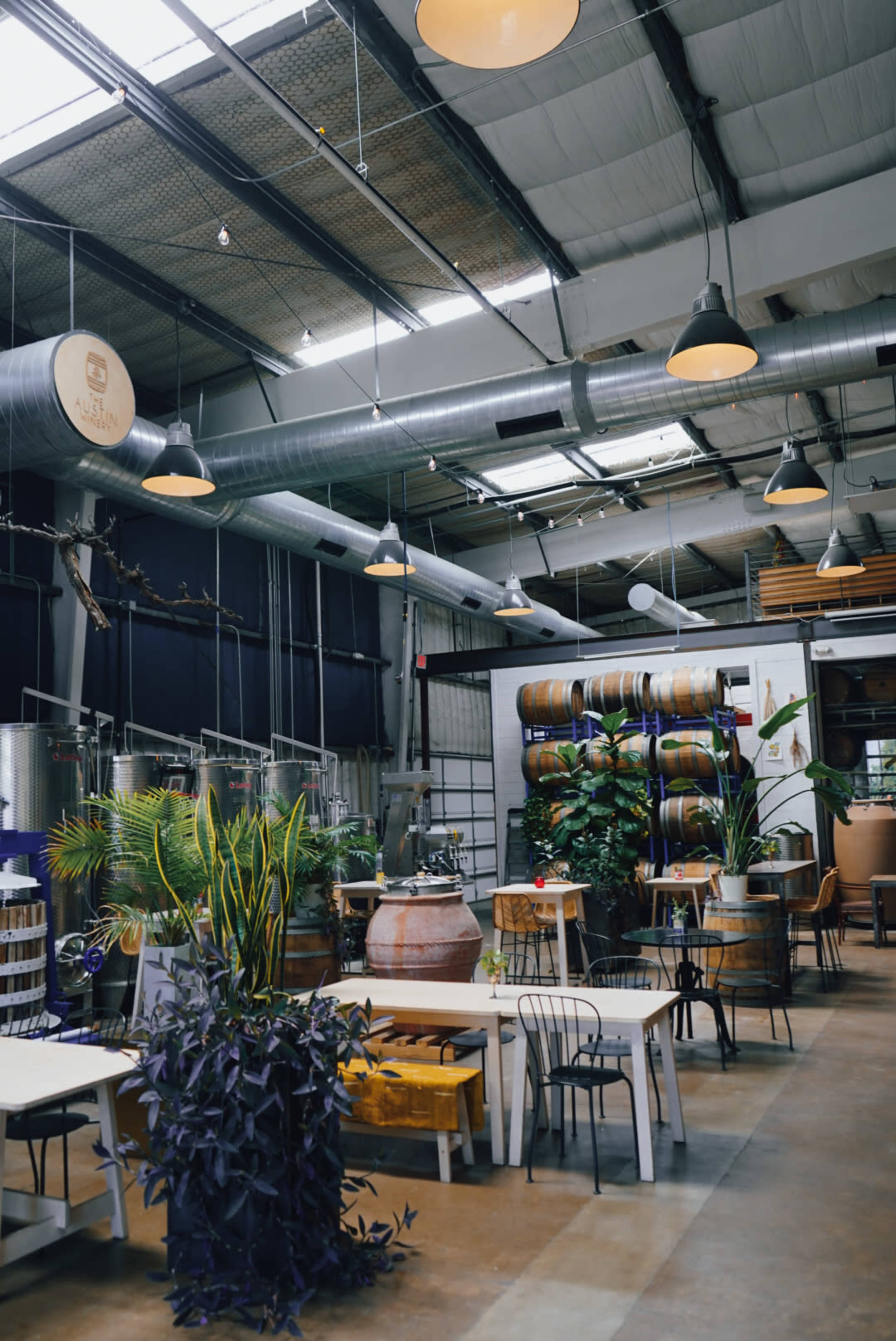 The image shows a spacious, industrial-style interior of a restaurant or café featuring wooden tables, large plants, and barrels.