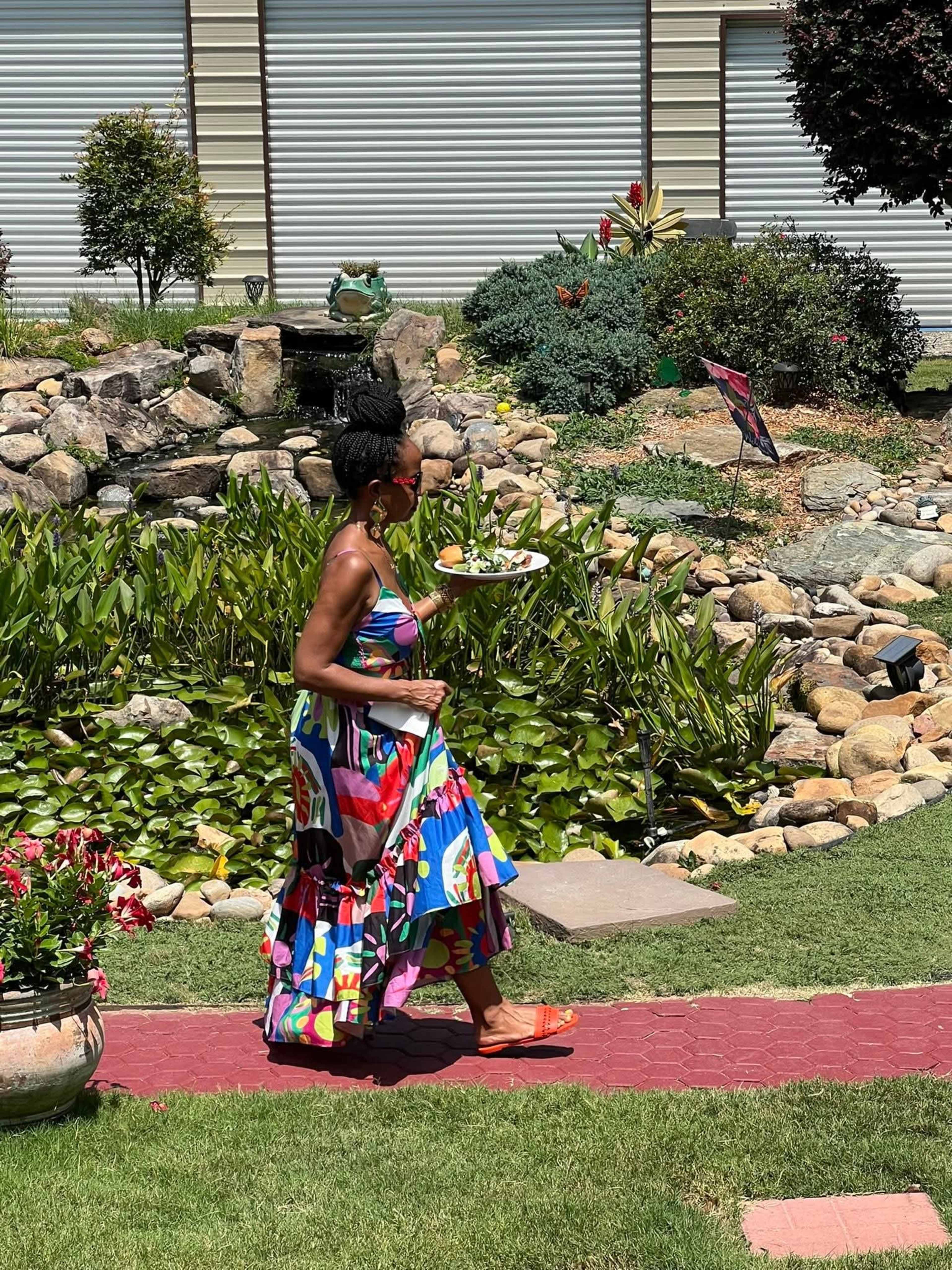 A woman wearing a colorful dress walks along a pathway while holding a plate in a garden filled with greenery and rocks.