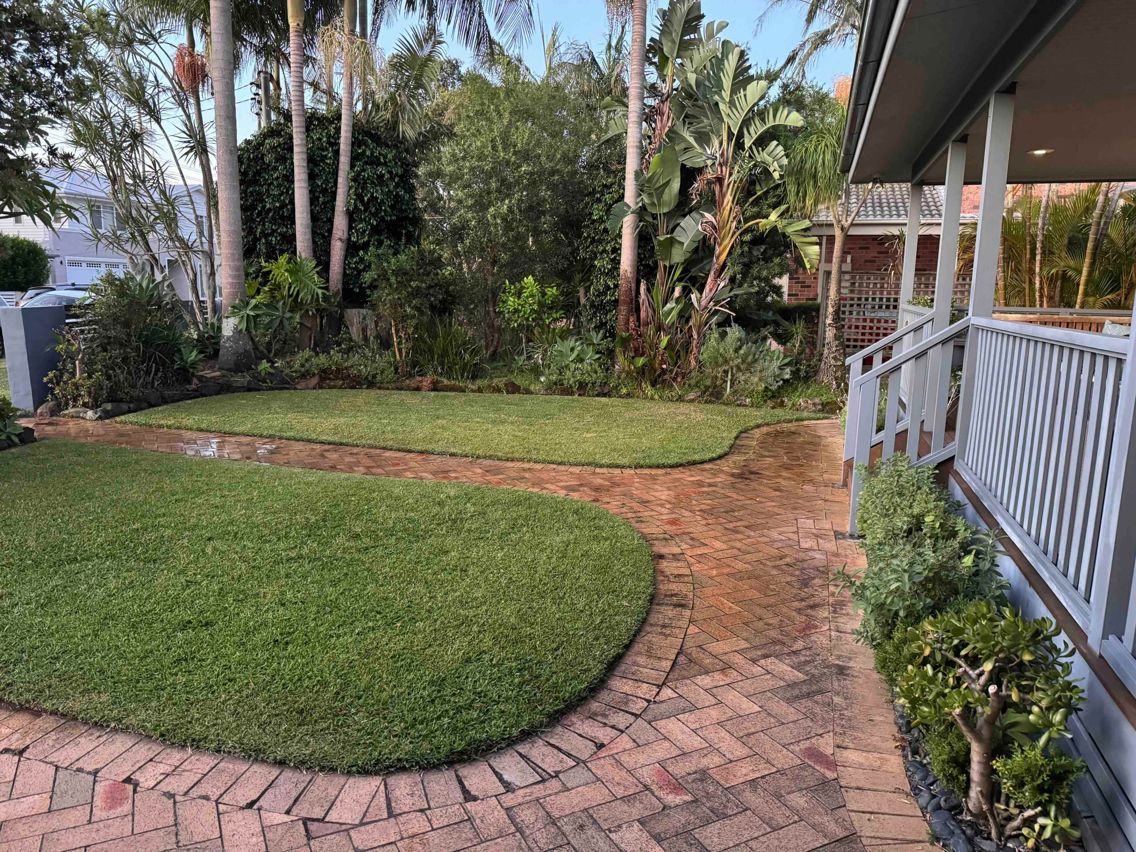 The image shows a landscaped yard featuring a curved brick pathway, green grass, and tropical plants surrounding the area.