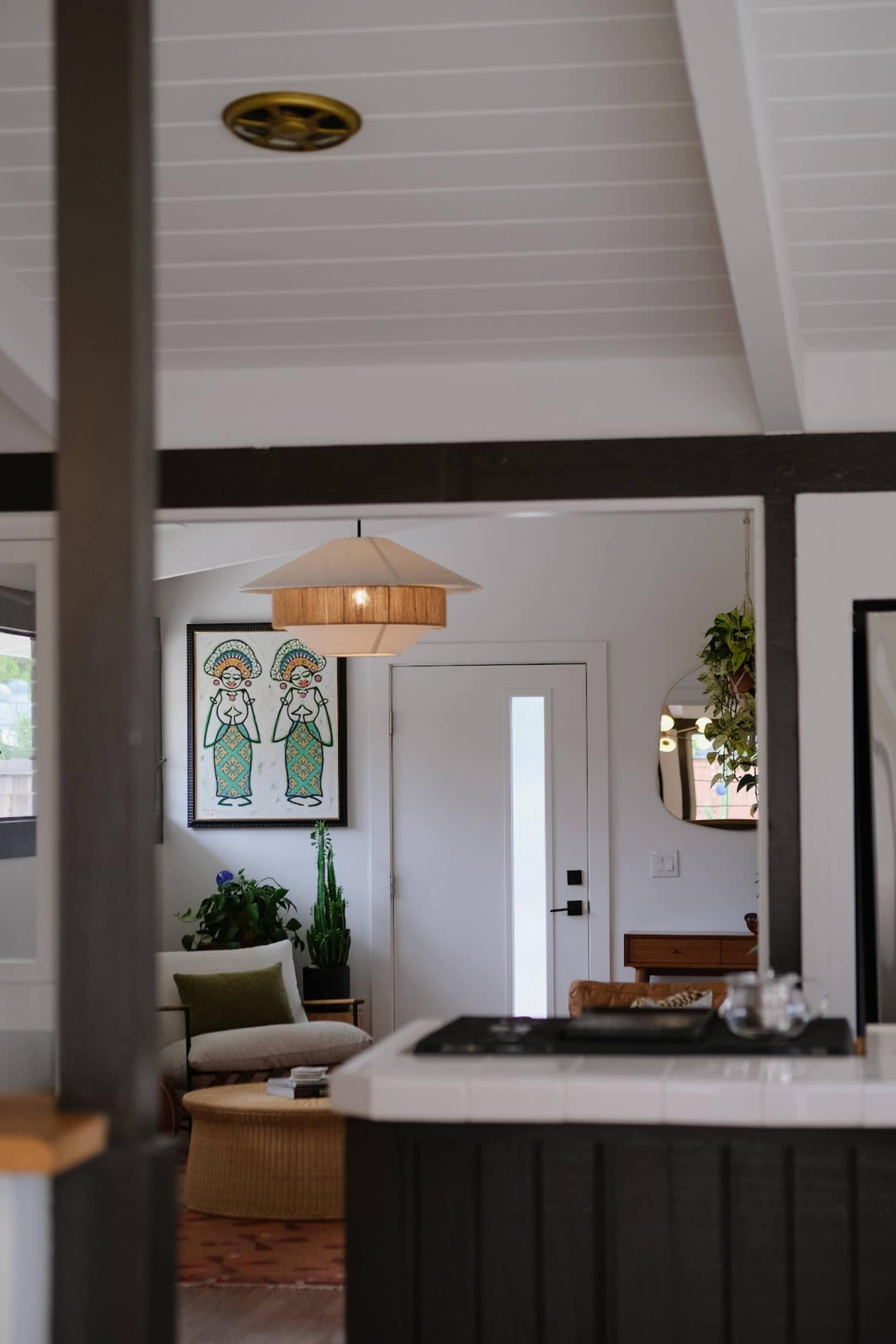The image shows a modern kitchen that opens into a living area, featuring a ceiling light fixture, framed artwork, and plants near a seating area.