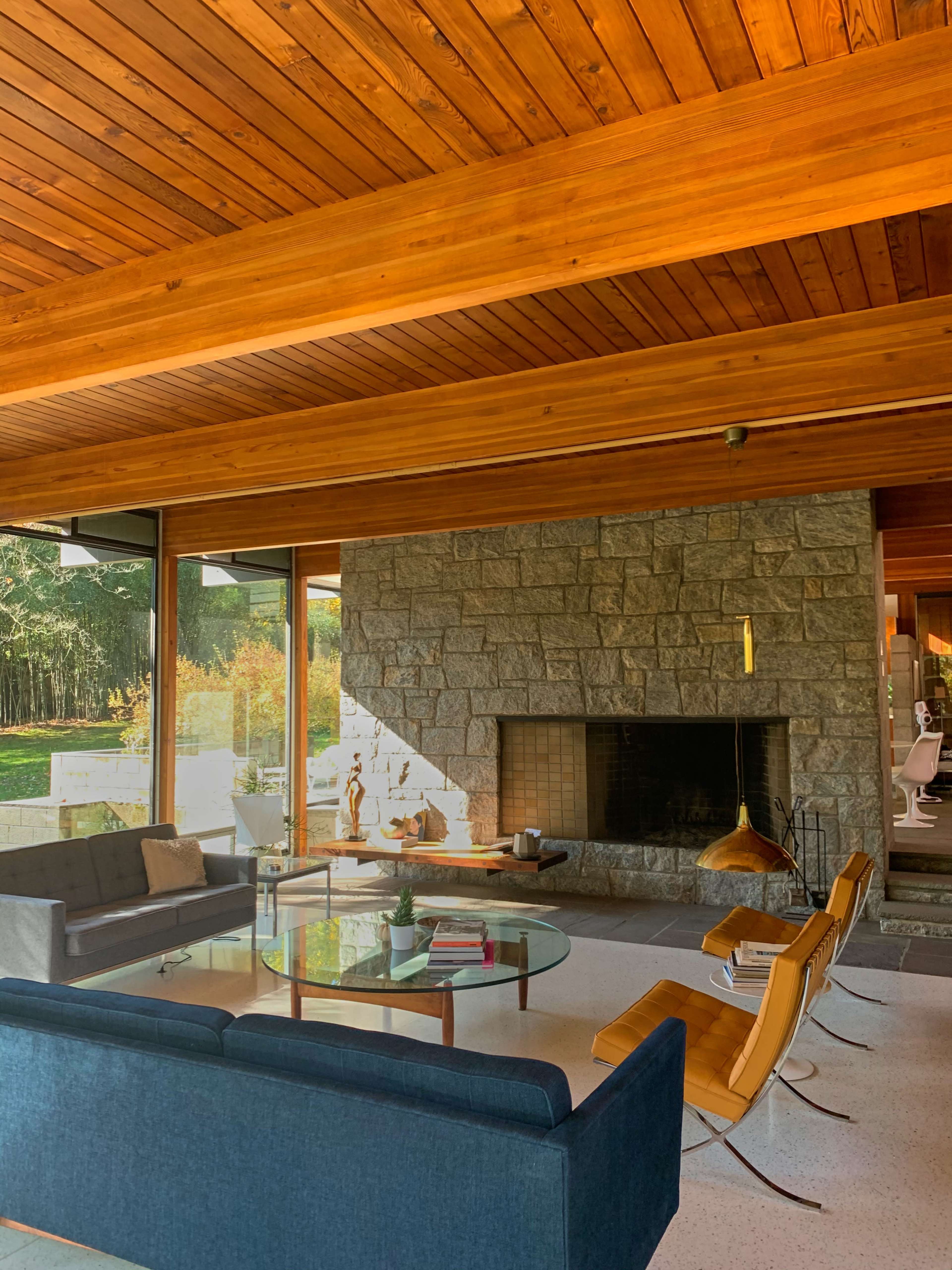 A modern living room features a stone fireplace, wooden ceilings, and a glass coffee table surrounded by minimalist furniture.