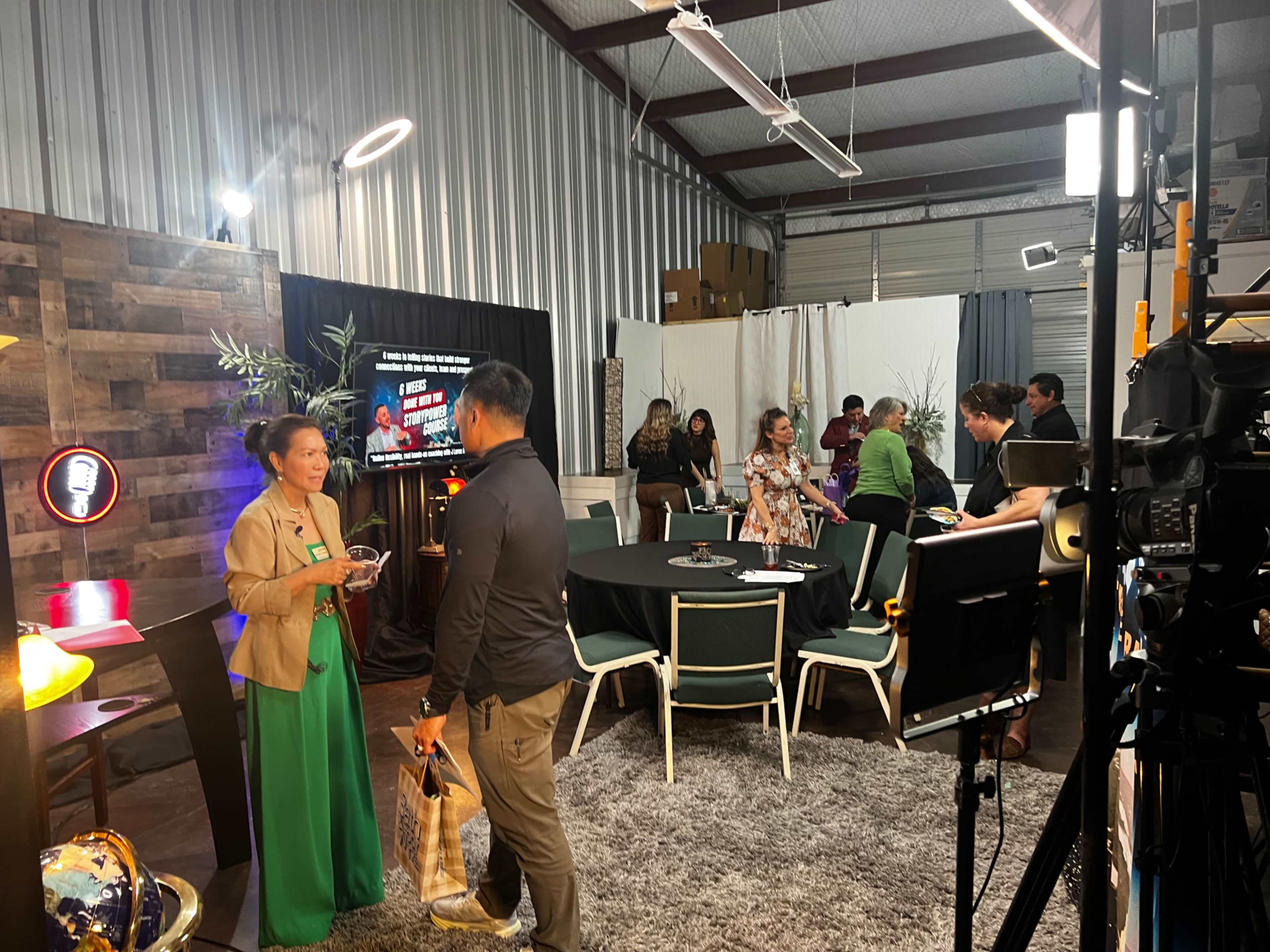 A group of people mingles in a well-lit event space featuring a decorated table, a screen displaying text, and various setup equipment.