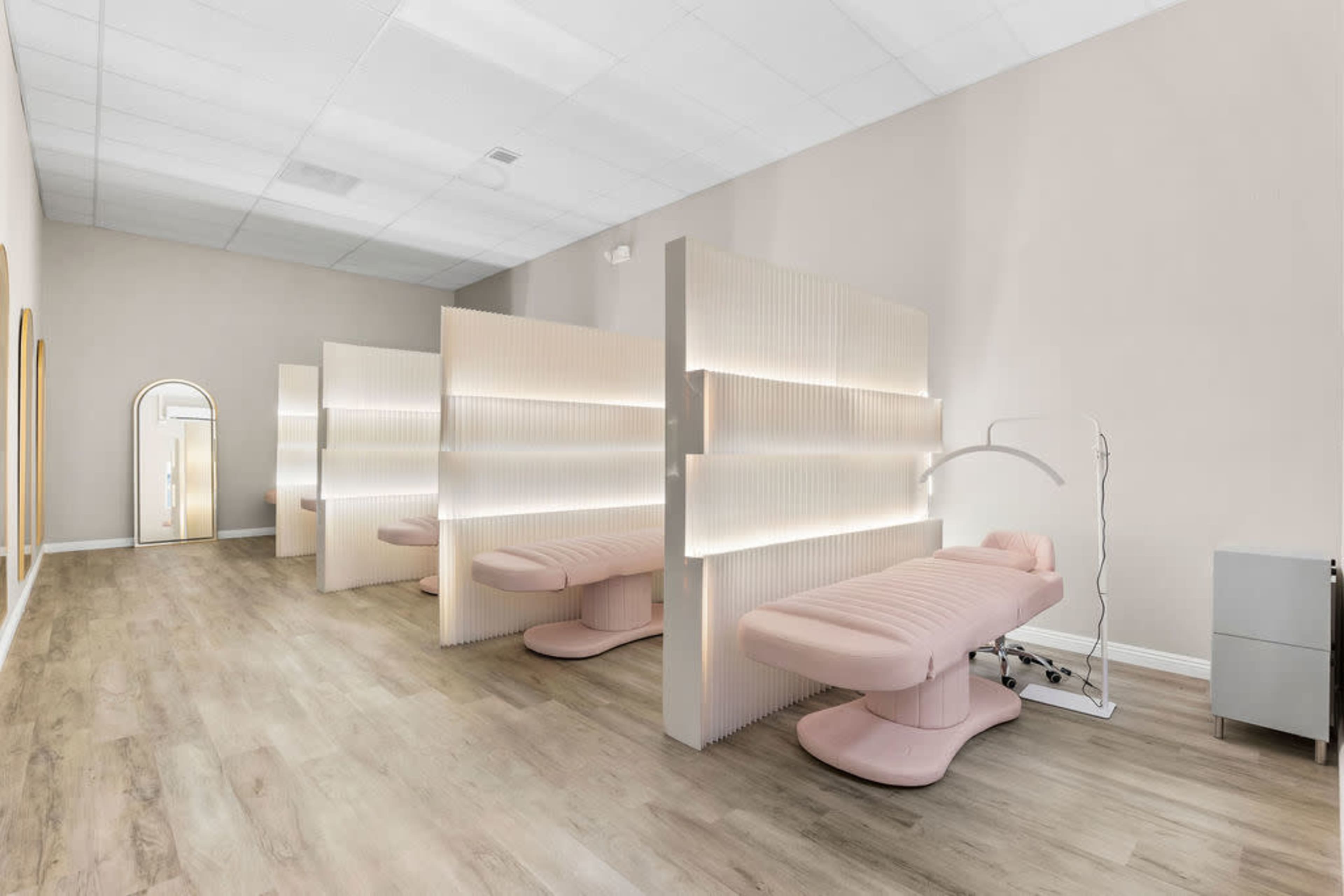 The image depicts a modern beauty salon interior featuring several pink treatment chairs separated by illuminated partitions on a light wood floor.