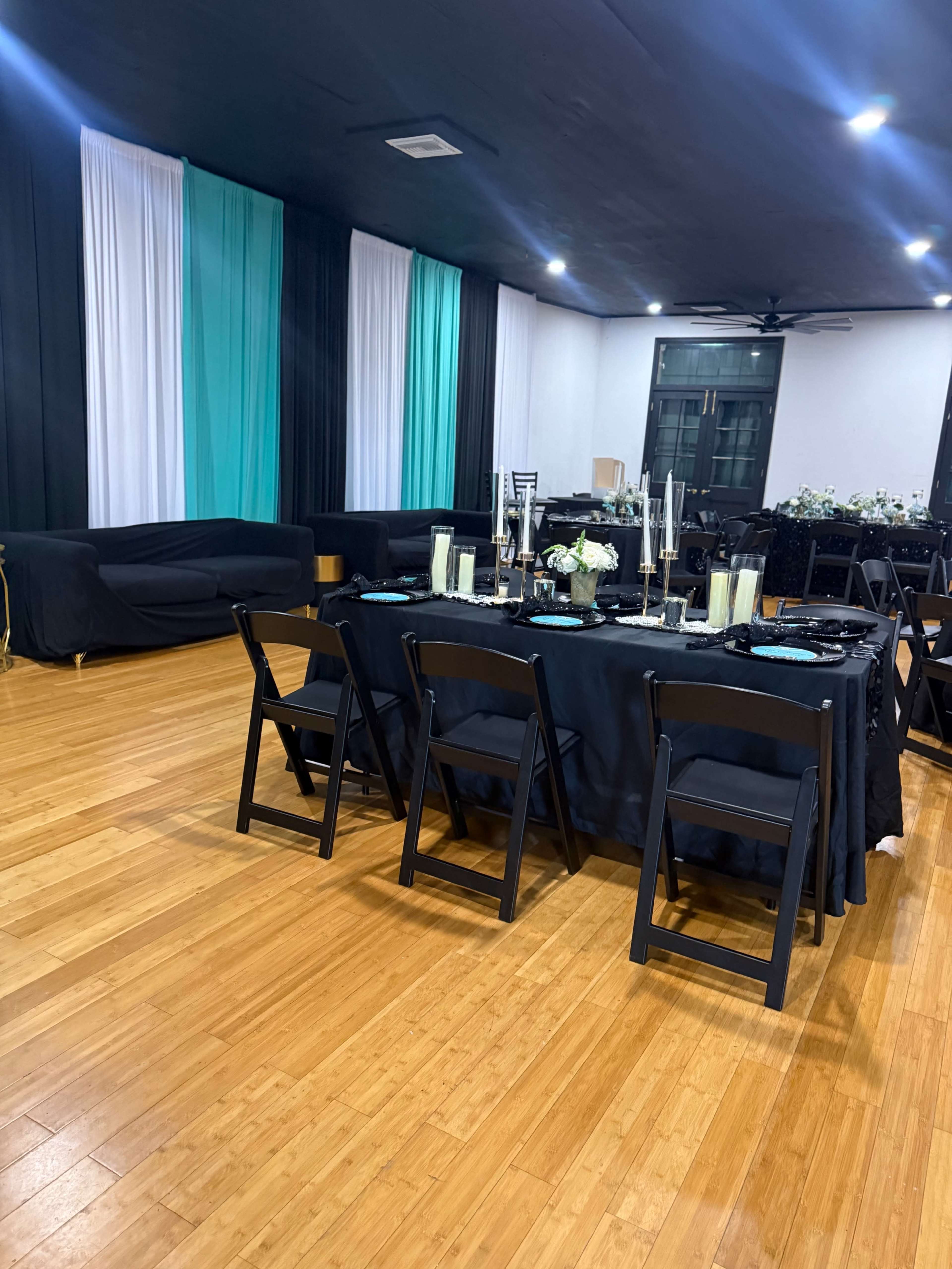 The image shows a decorated event space featuring a black table set with candles and flowers, surrounded by black folding chairs, and draped in teal and white fabric on the walls.