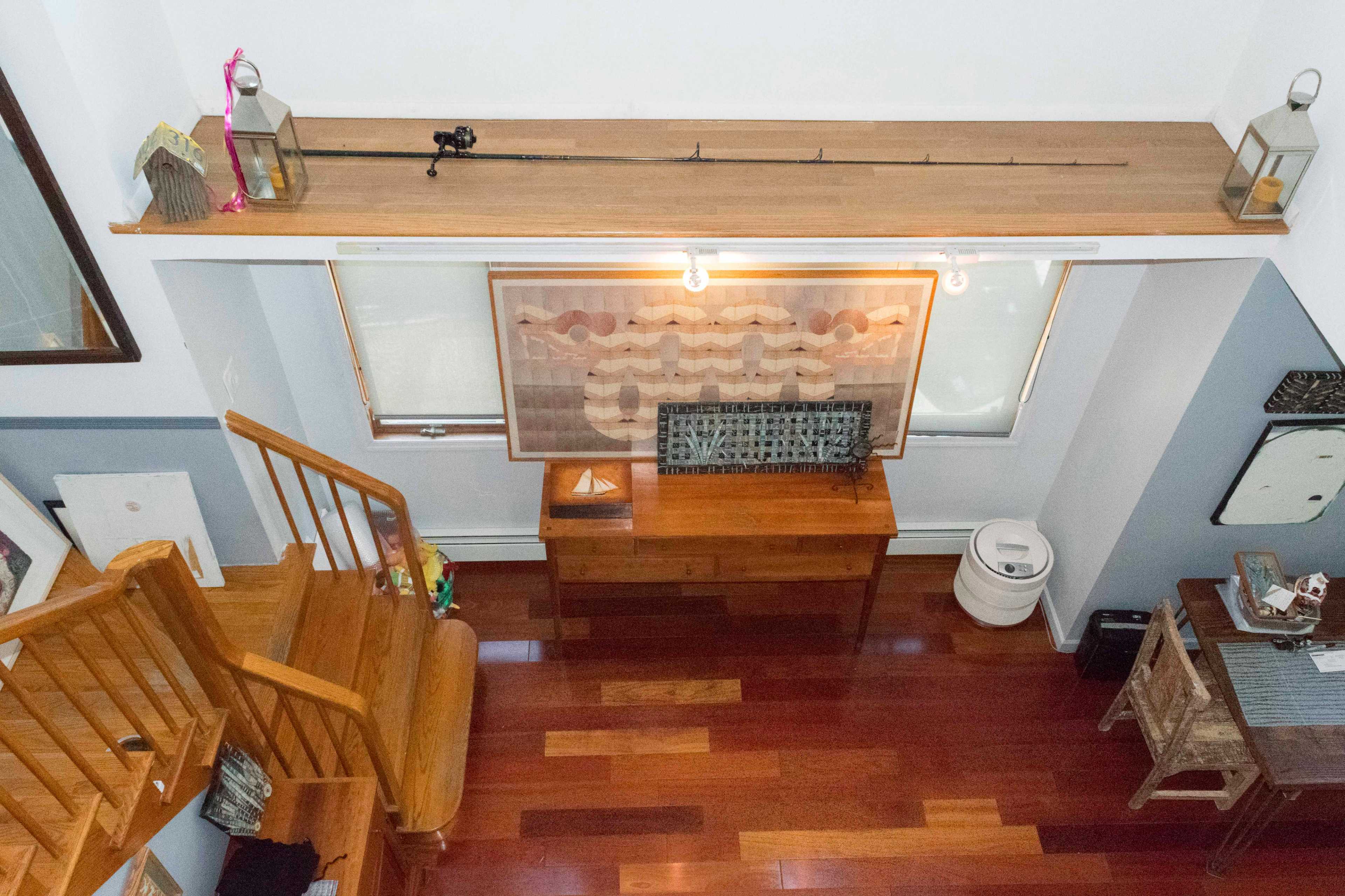 The image shows a spacious room from an elevated angle featuring wooden flooring, a wooden desk with a patterned artwork above it, and a staircase on the left side.