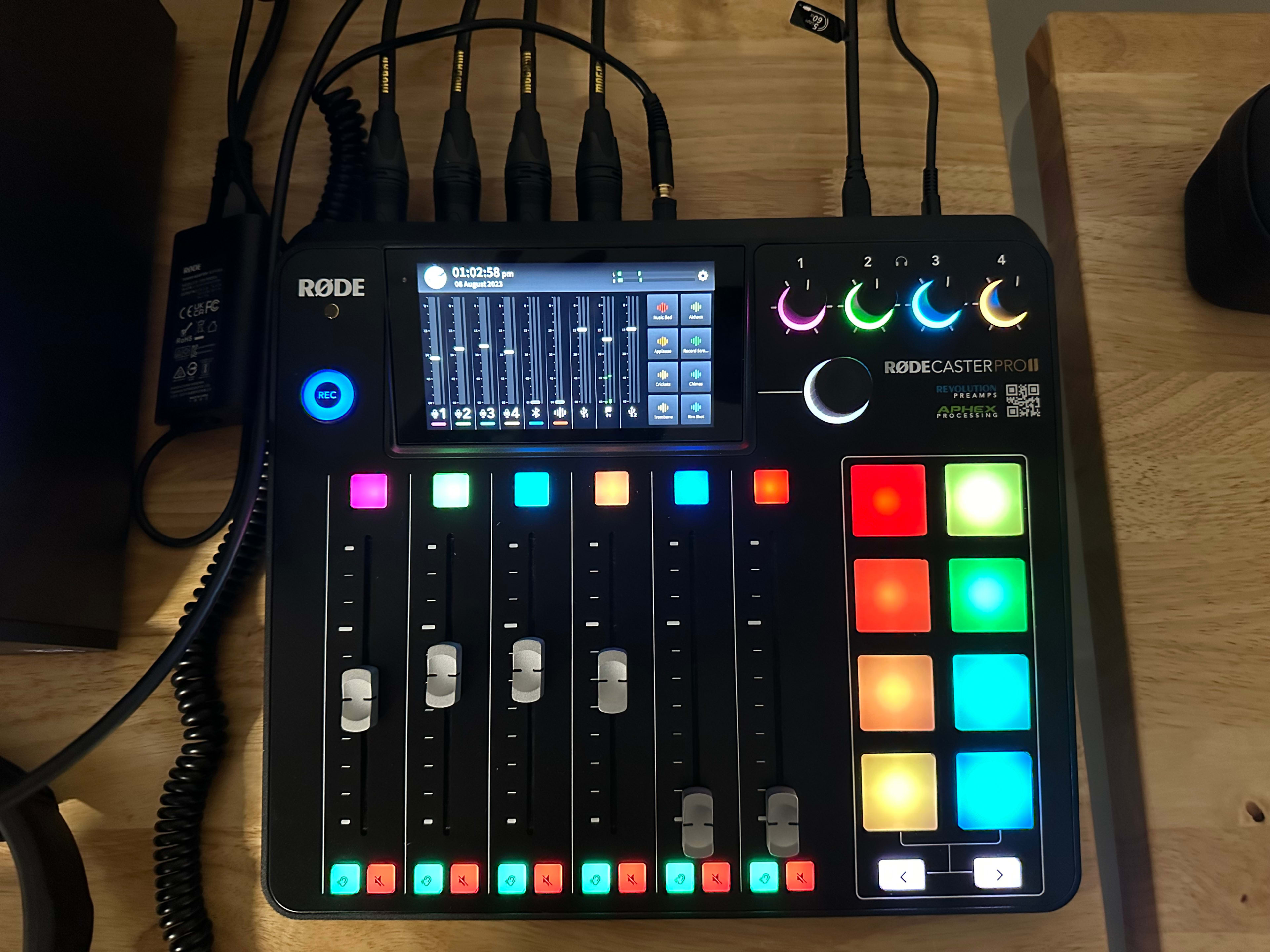 The image shows a Rodecaster Pro II audio mixing console featuring a touchscreen display, multiple colorful buttons, and faders.
