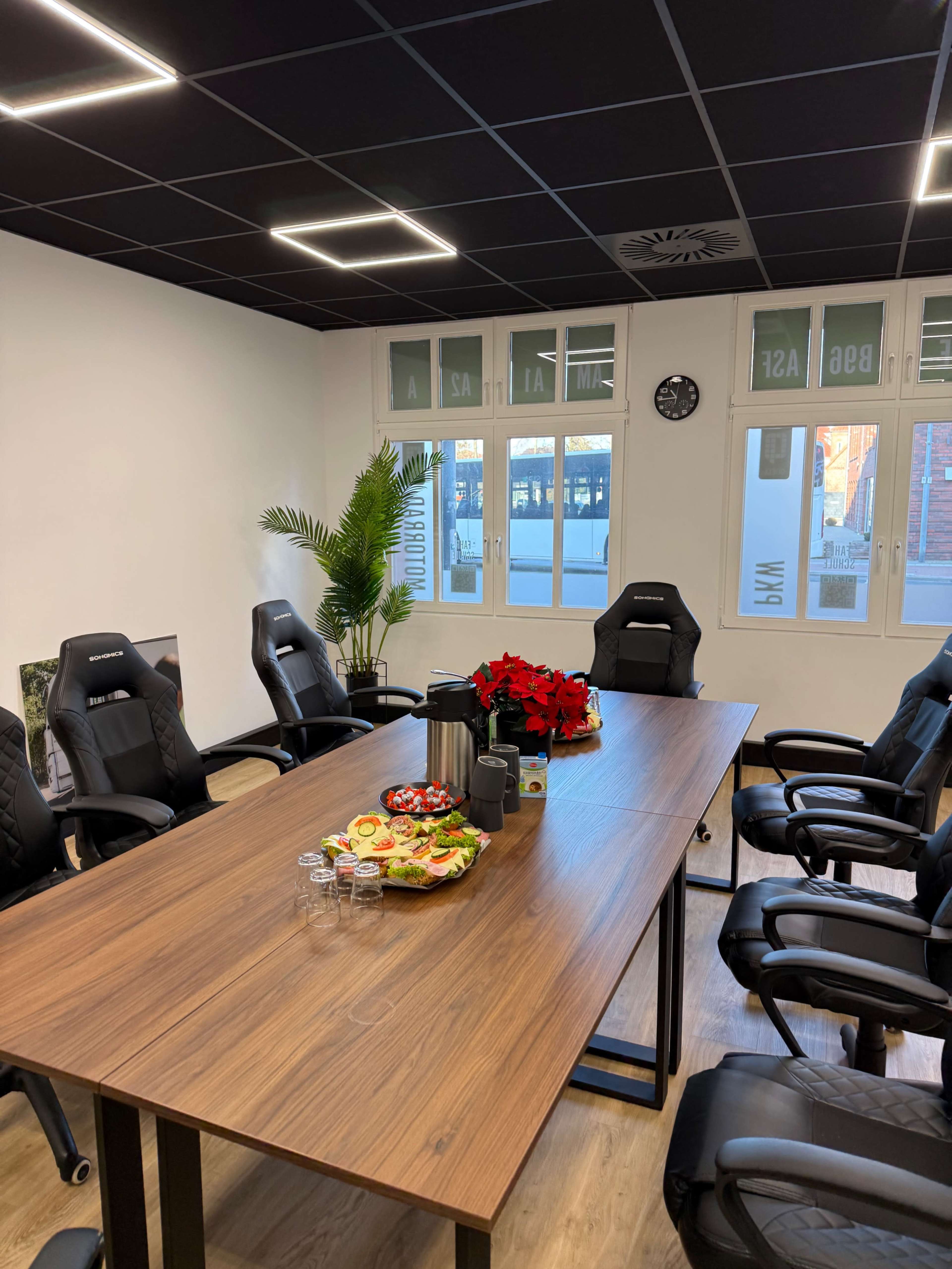 The image shows a modern conference room with a large wooden table surrounded by ergonomic chairs, decorated with a plant and a tray of snacks.