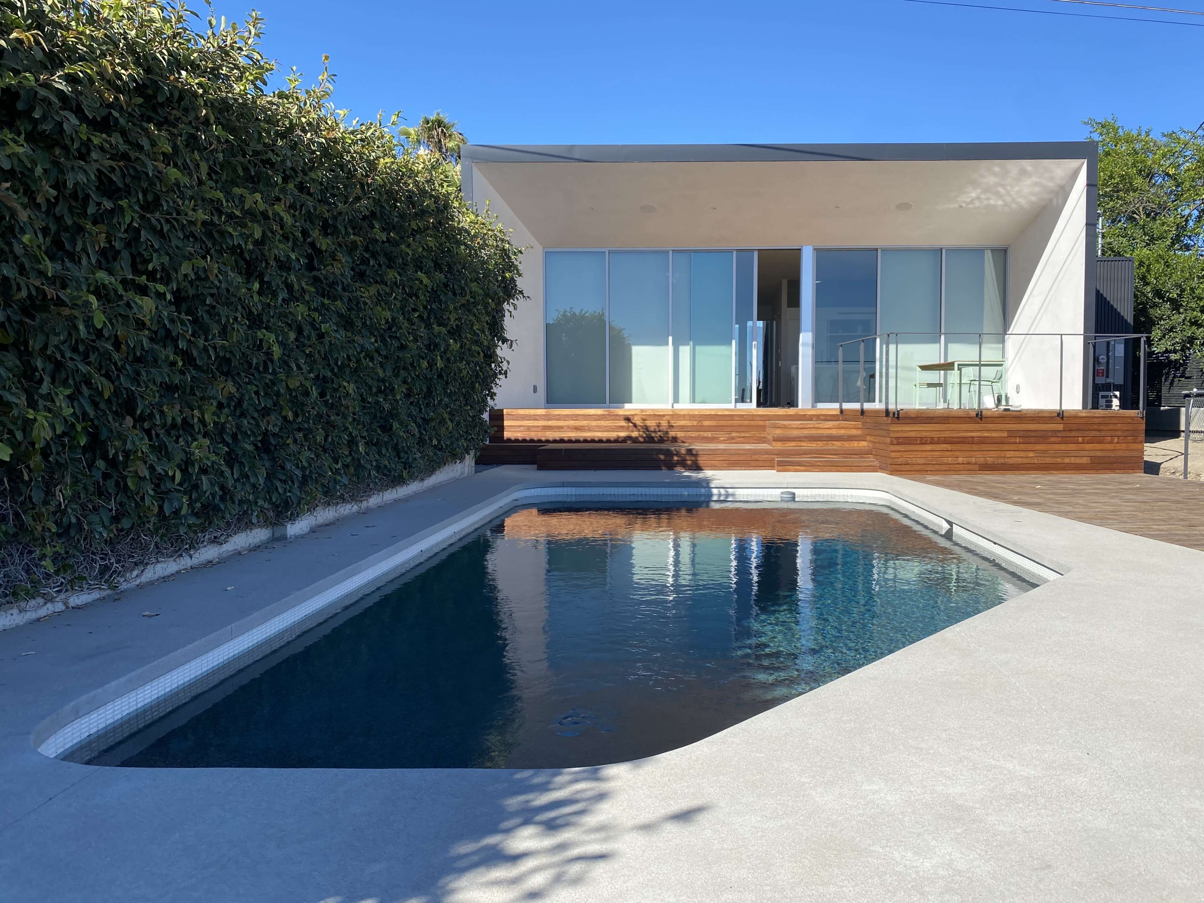 A modern house with large glass windows overlooks a rectangular swimming pool surrounded by a concrete patio and a tall hedge.