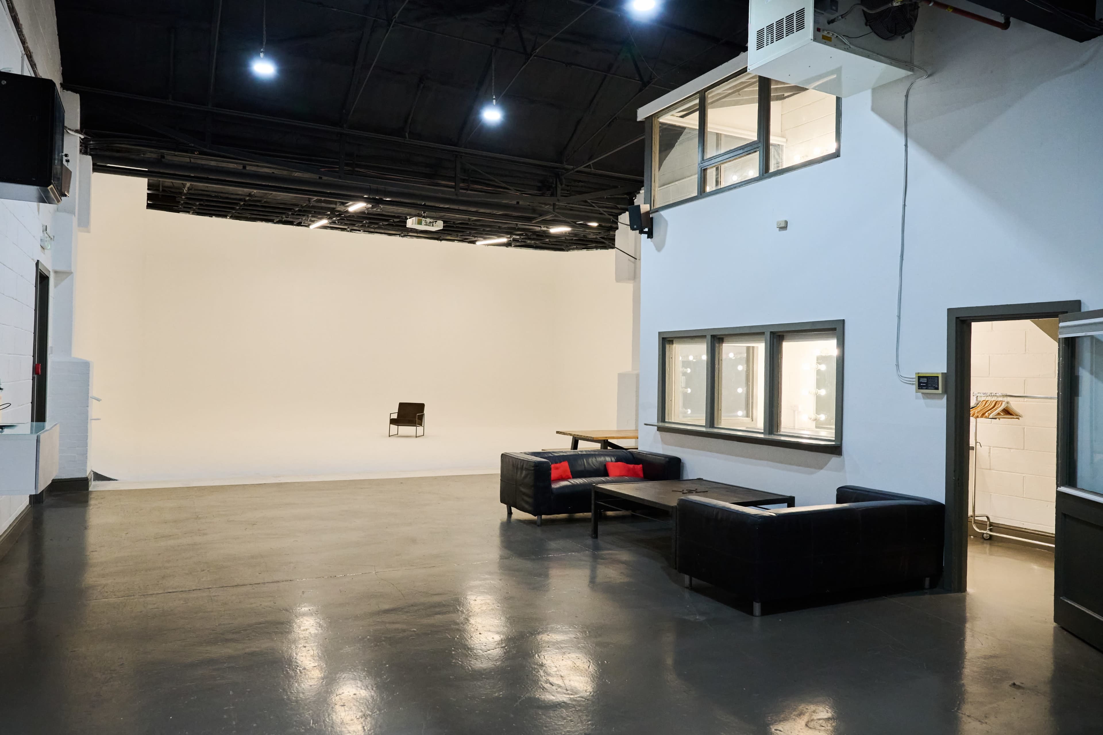 The image shows a spacious studio with a white backdrop, featuring a single chair in the center and a waiting area with black couches and large windows.