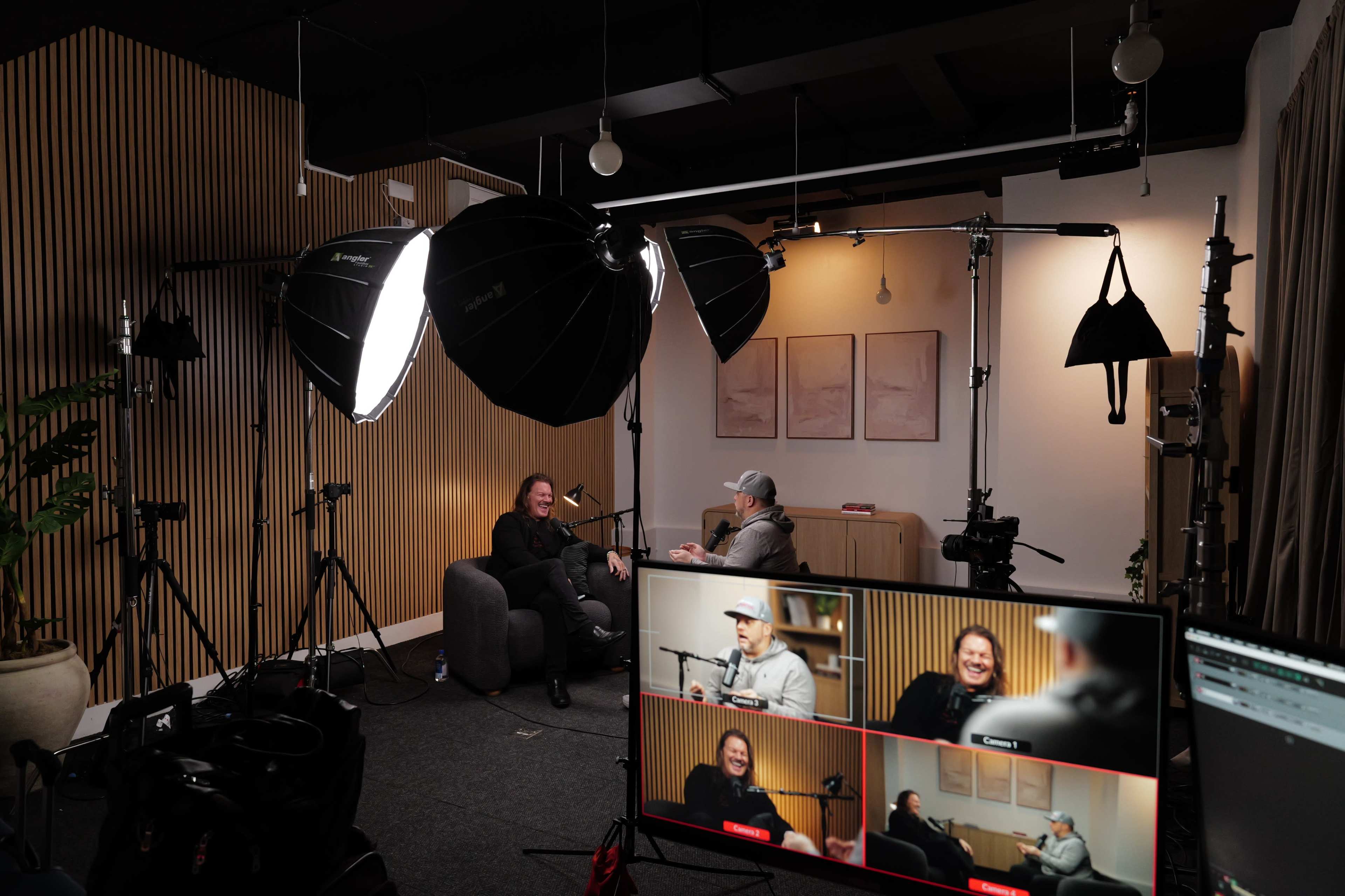 The image shows a well-set studio with two people seated on a gray couch, bright lights focused on them, and multiple cameras positioned around, while a monitor displays their conversation from different angles.