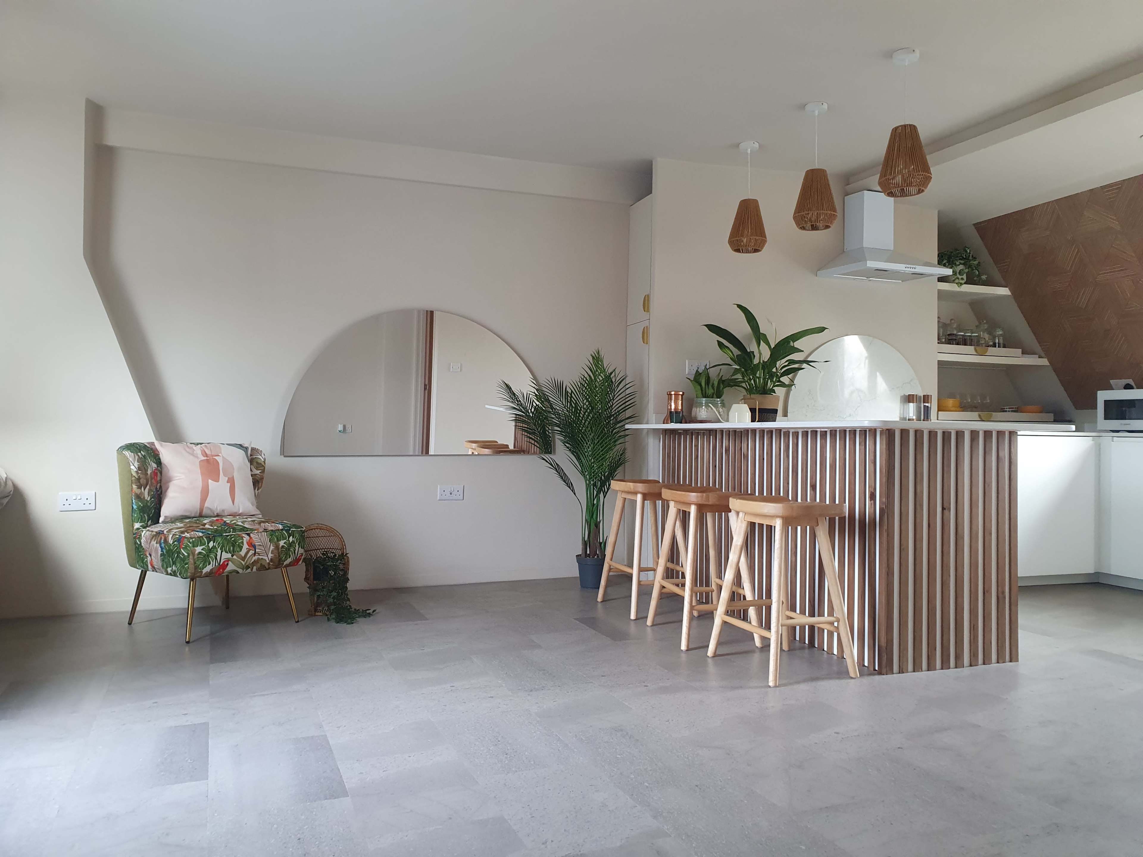 A modern kitchen and living area with a wooden countertop, three stools, and a tropical-themed chair beside a round mirror.