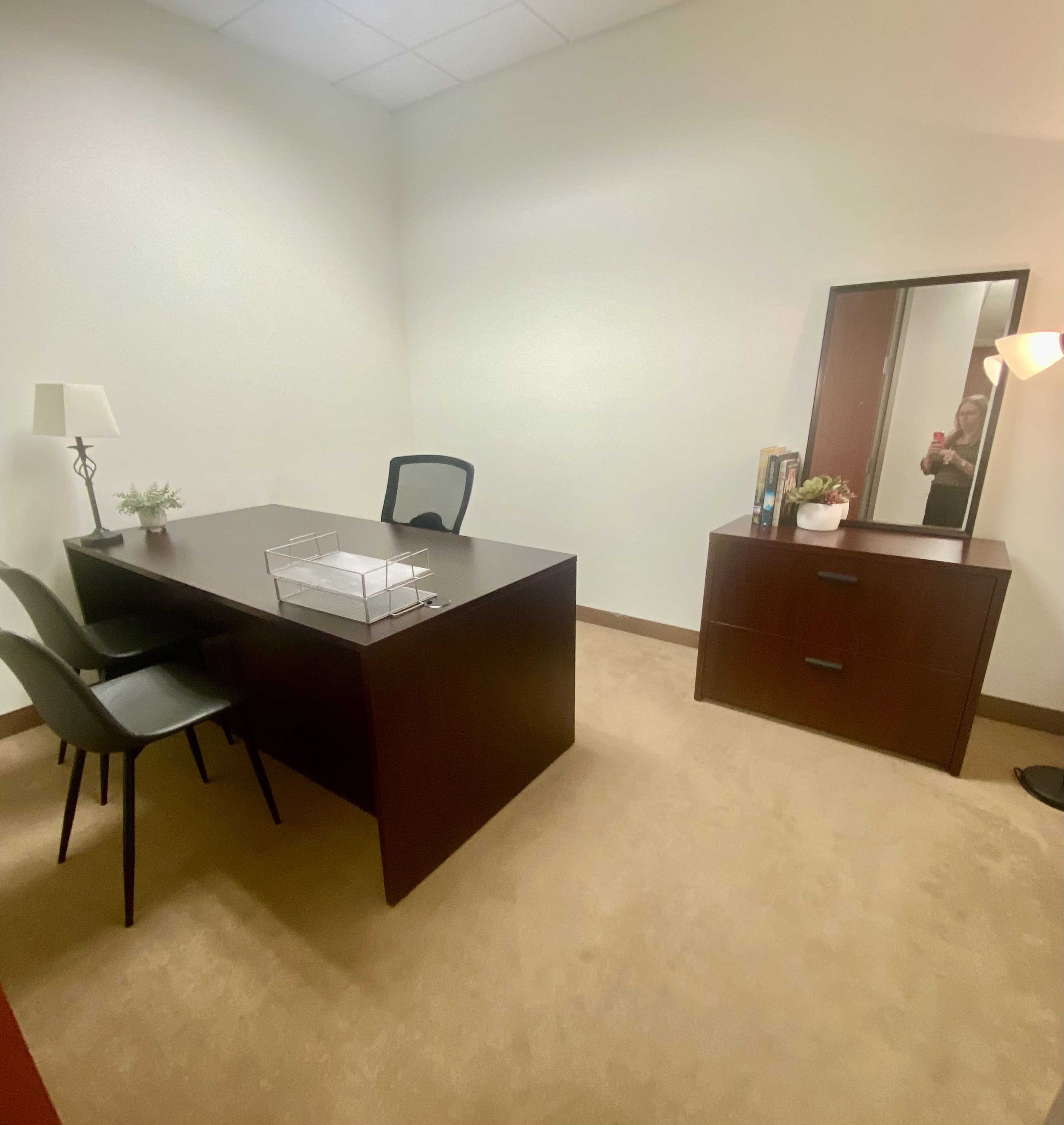 The image shows a minimalist office space with a dark wood desk, two chairs, a dresser, and a mirror against a neutral-colored wall.