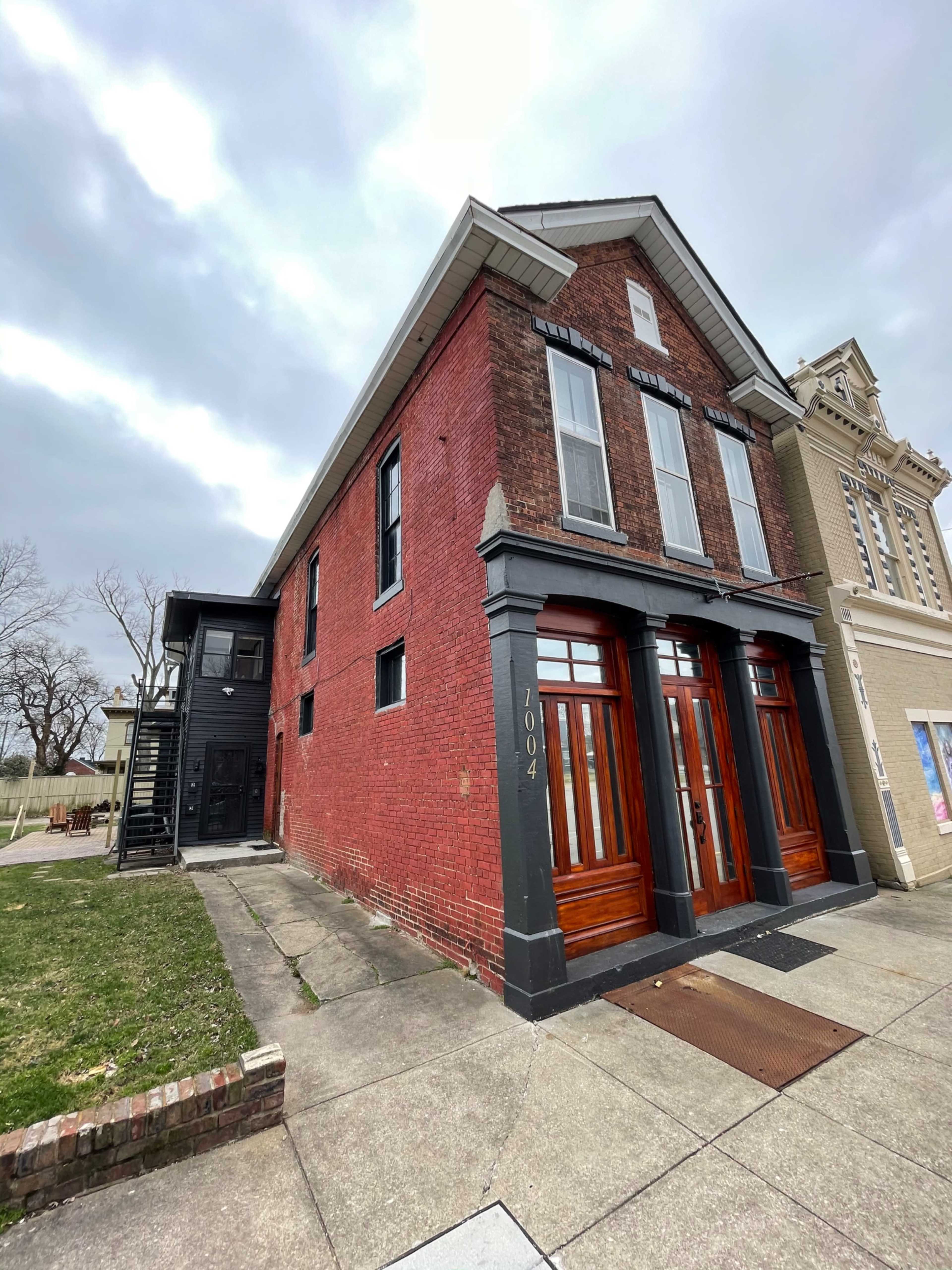 A two-story red brick building with large wooden doors, situated on a corner lot adjacent to another building.
