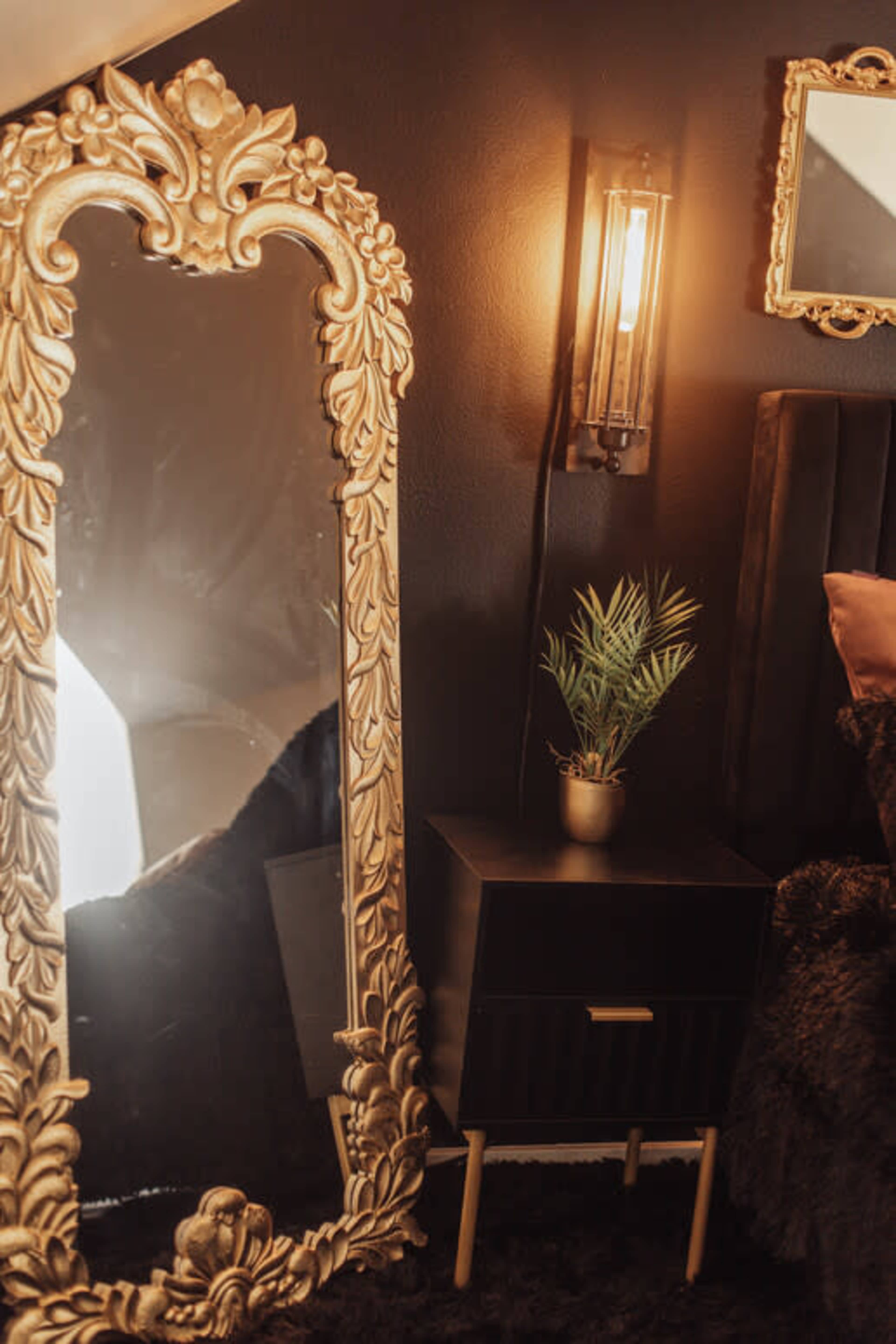 The image features a decorative gold-framed mirror beside a black nightstand with a small potted plant and a wall-mounted light fixture.