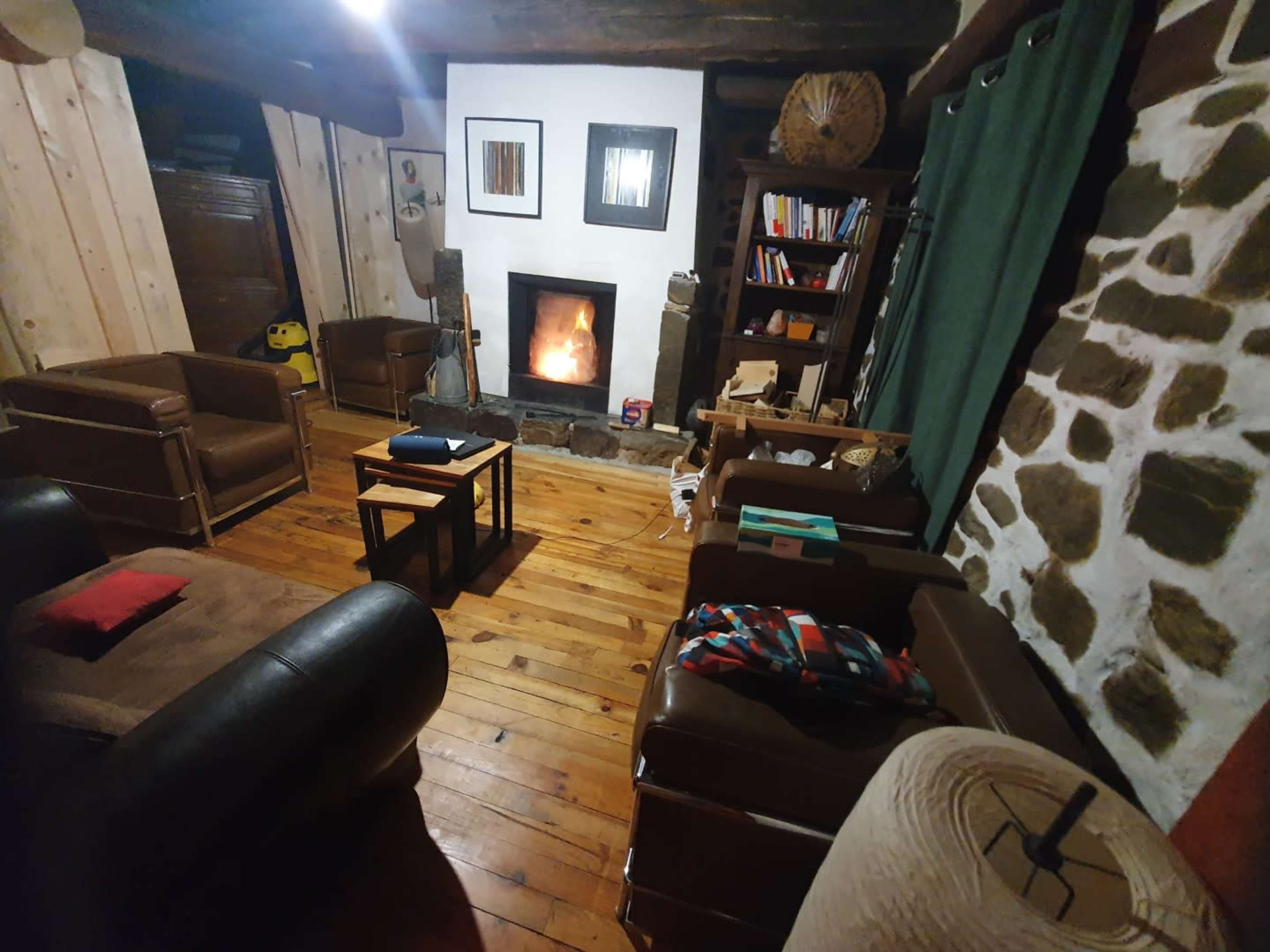 The image shows a cozy room with wooden flooring, stone walls, a fireplace, and seating arrangements including brown chairs and a small wooden table.