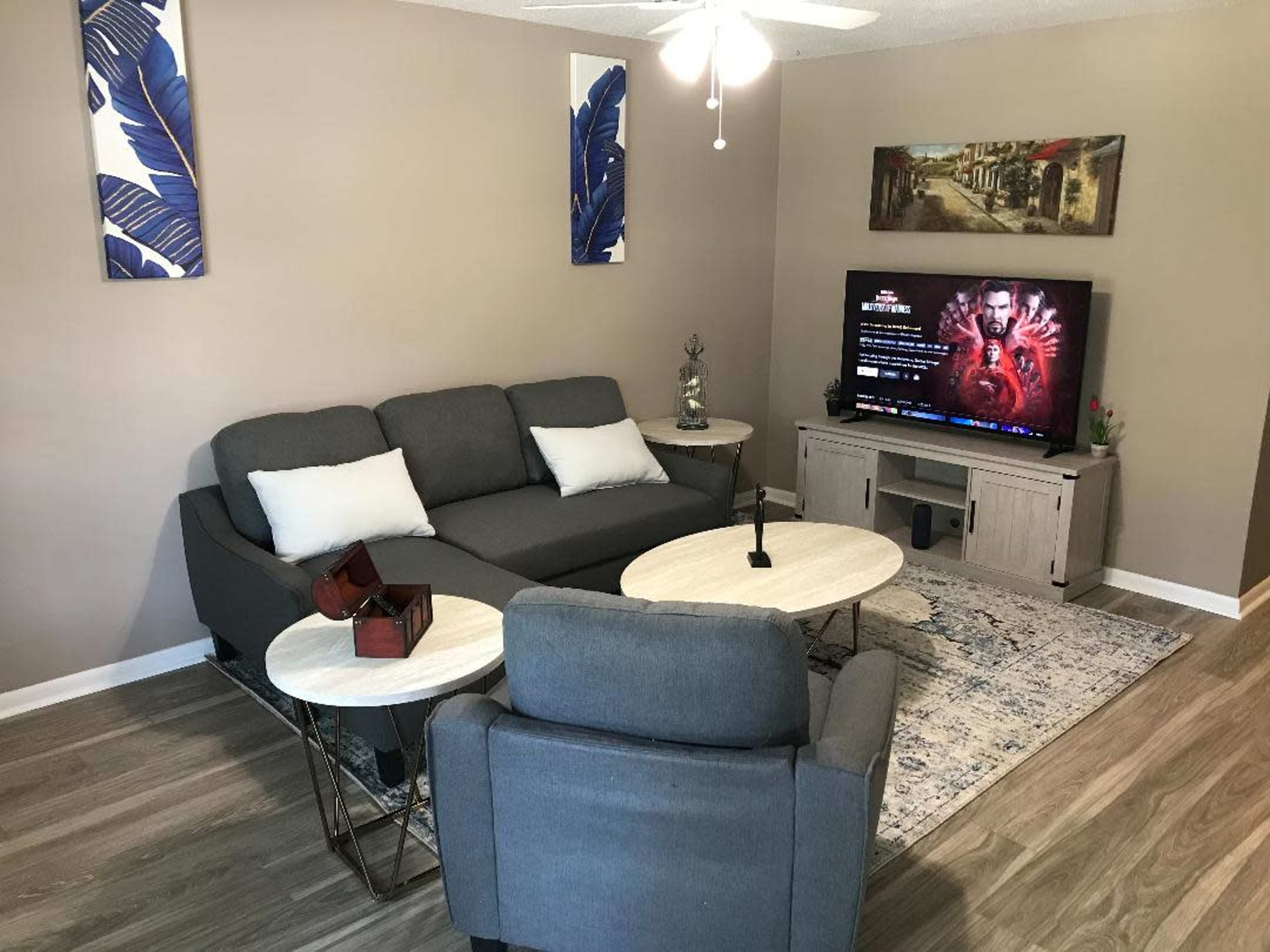 A living room with a gray sectional sofa, two small coffee tables, a television displaying content, and decorative wall art.