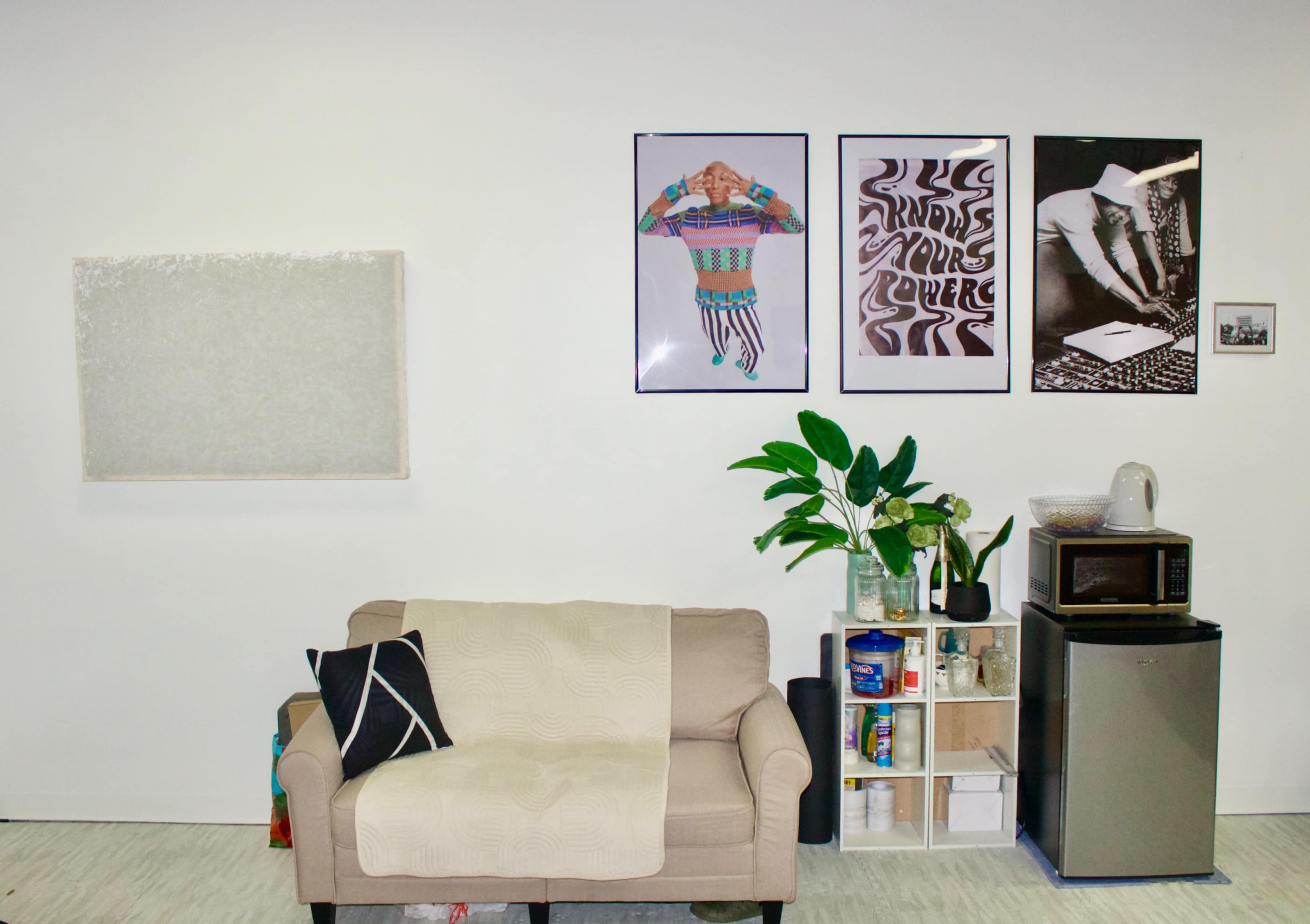 The image shows a living space featuring a beige sofa, a small shelf with storage items, a microwave, and several framed artworks on the wall.