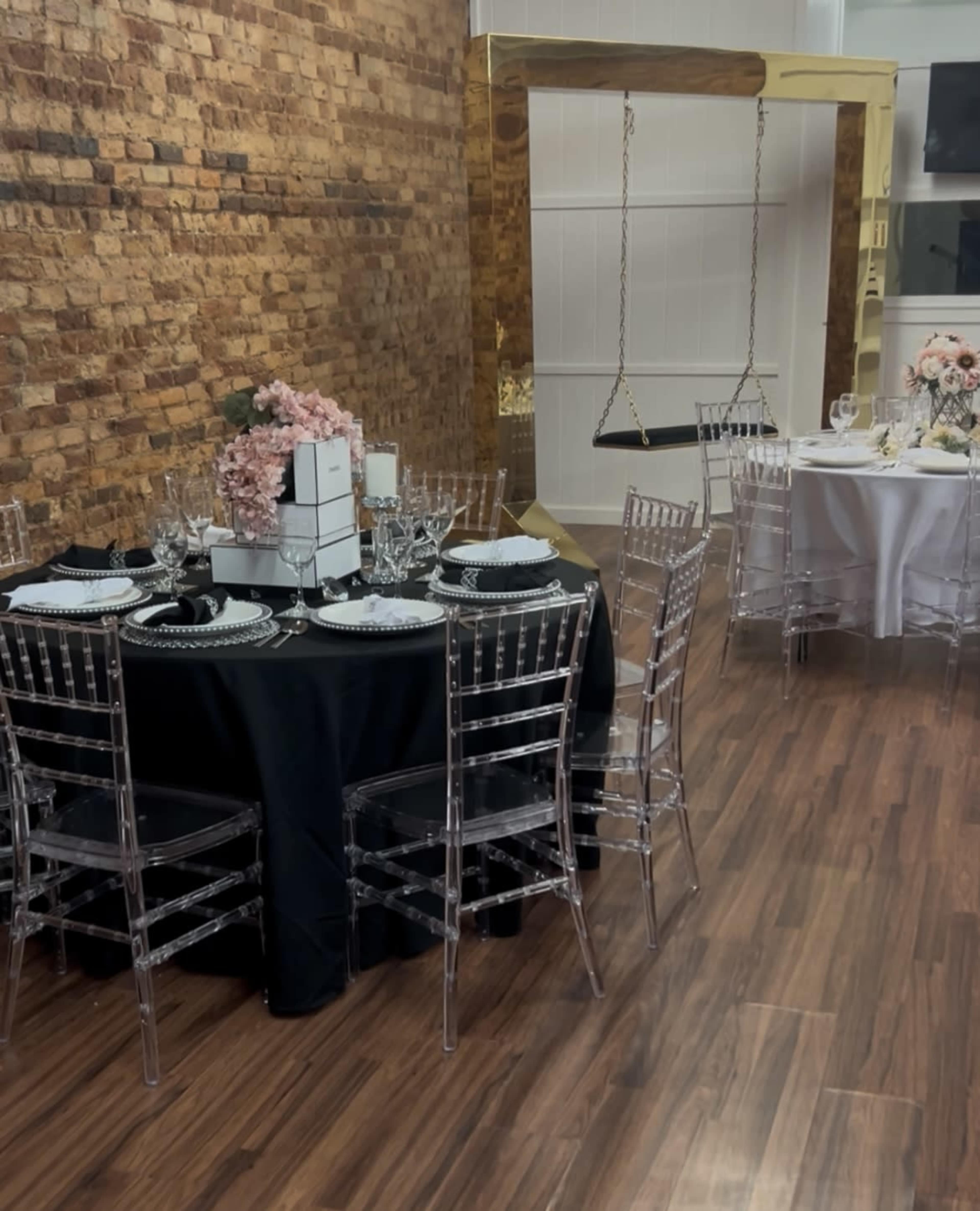 The image shows a room with a wooden floor featuring two elegantly set dining tables, one with a black tablecloth and clear chairs, and another round table with a white tablecloth and floral centerpieces.