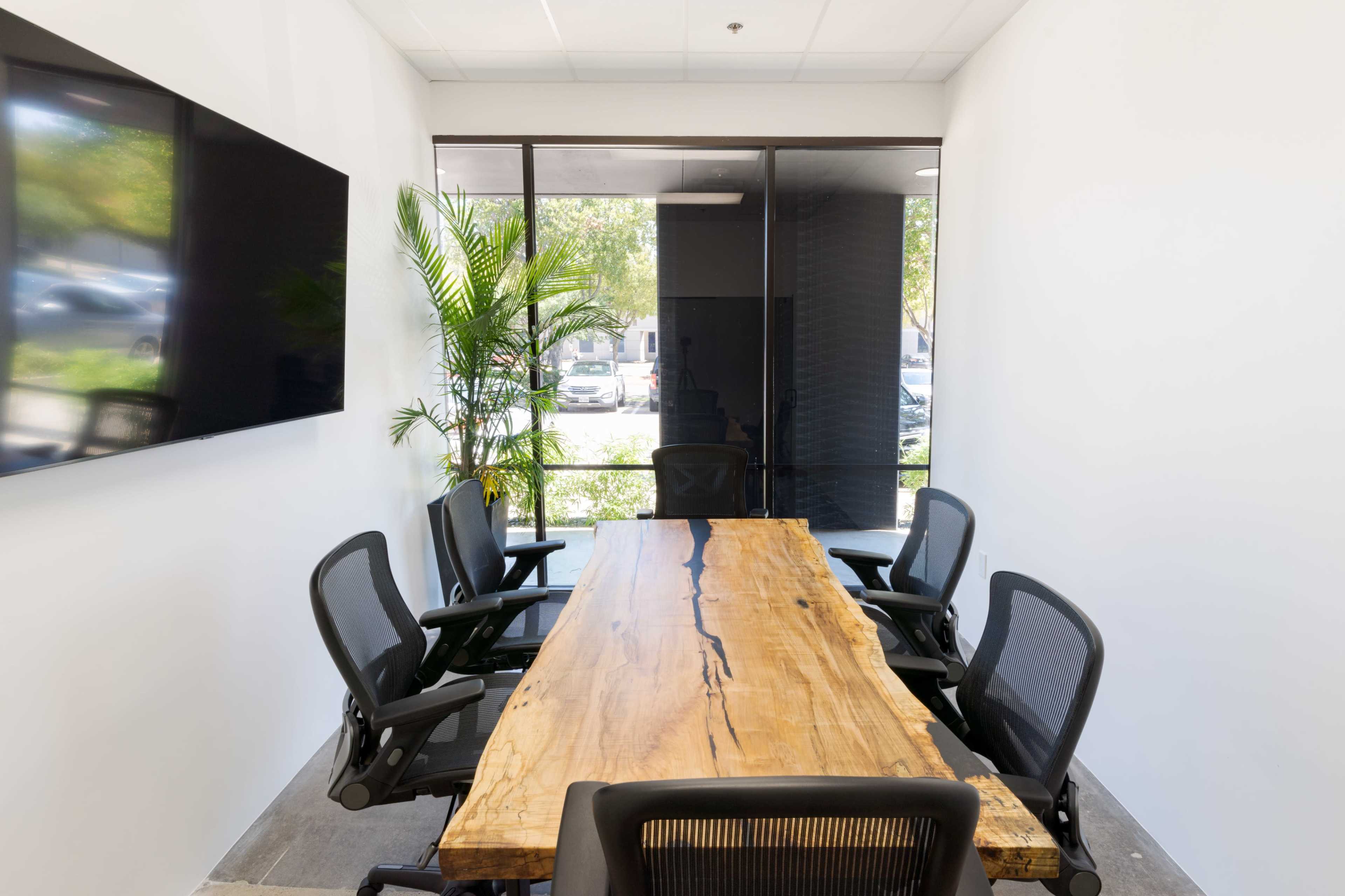 A modern conference room features a long wooden table surrounded by six black office chairs, with a large screen on one wall and sunlight streaming through large windows.