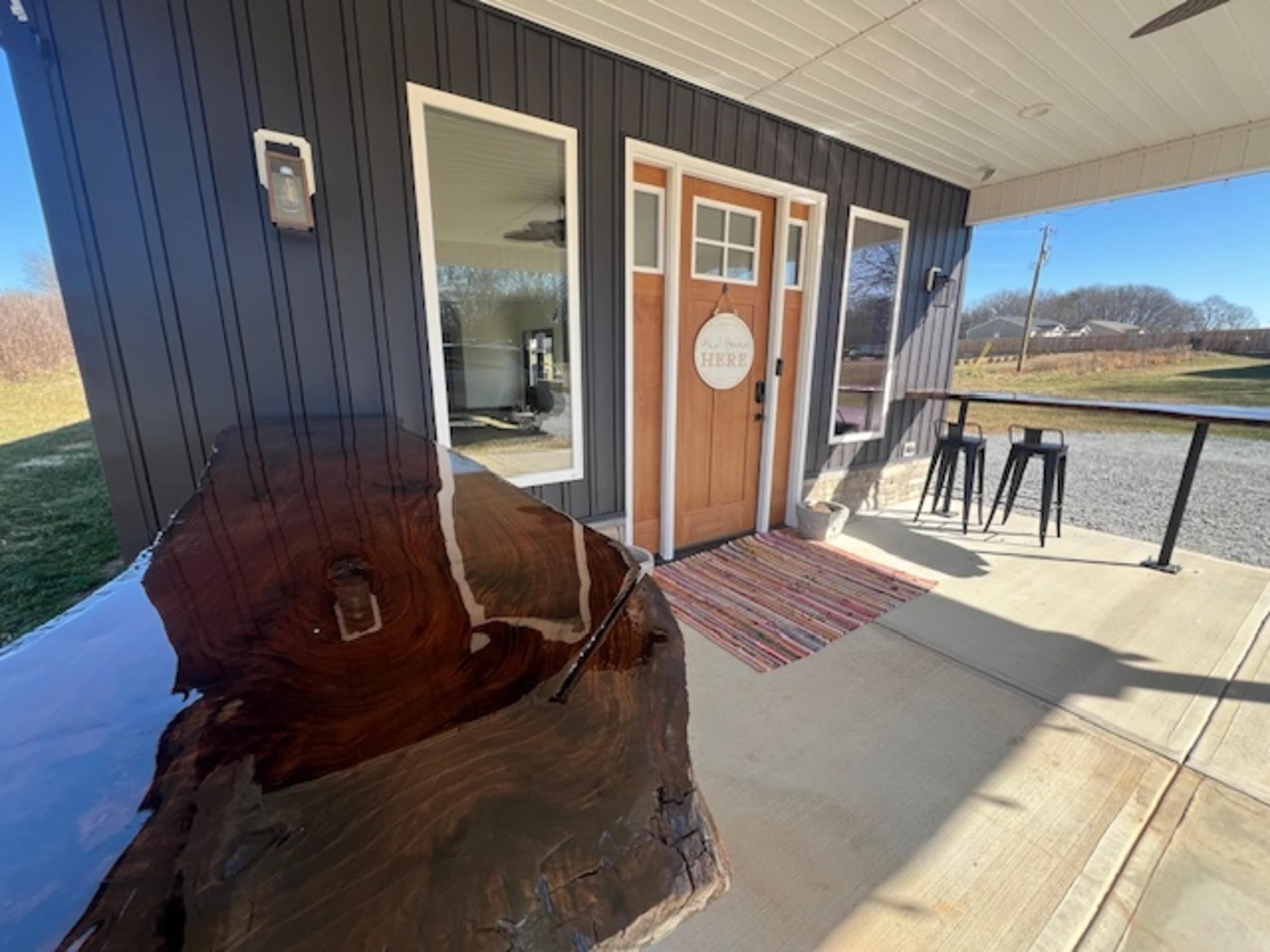 A wooden table is positioned beside the entrance of a modern building with a sign that reads "HERE," featuring large windows and a porch area.