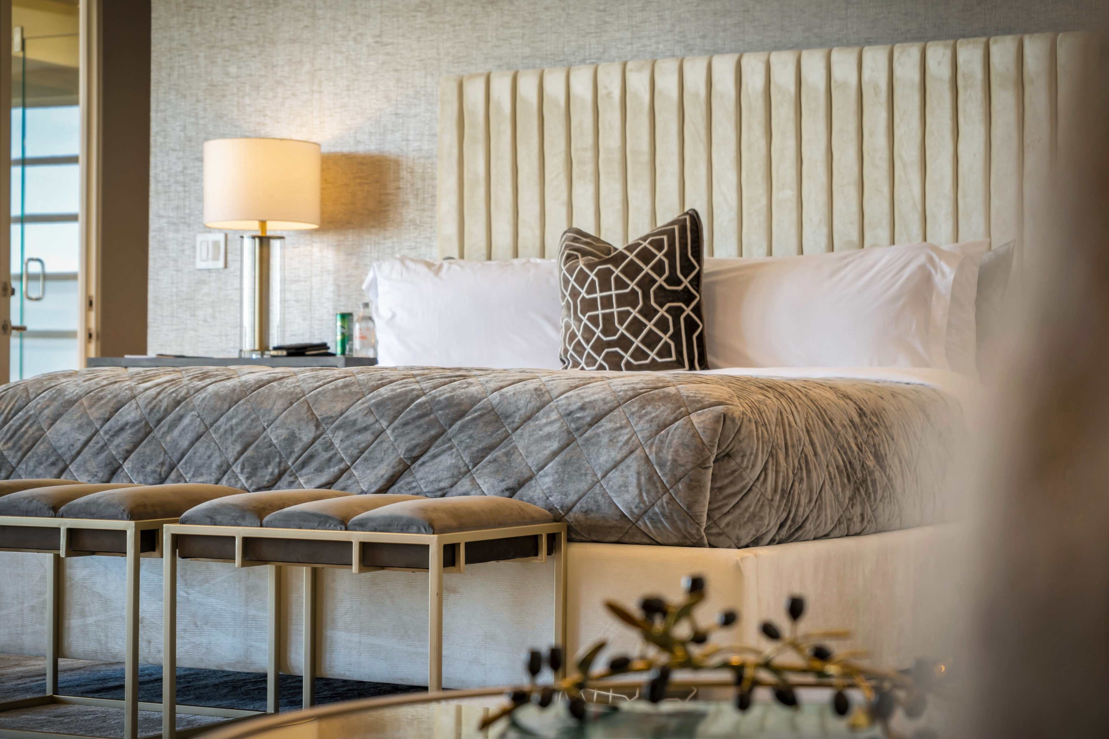 The image shows a neatly arranged hotel bedroom featuring a plush bed with a gray coverlet and decorative pillows, flanked by two upholstered benches.