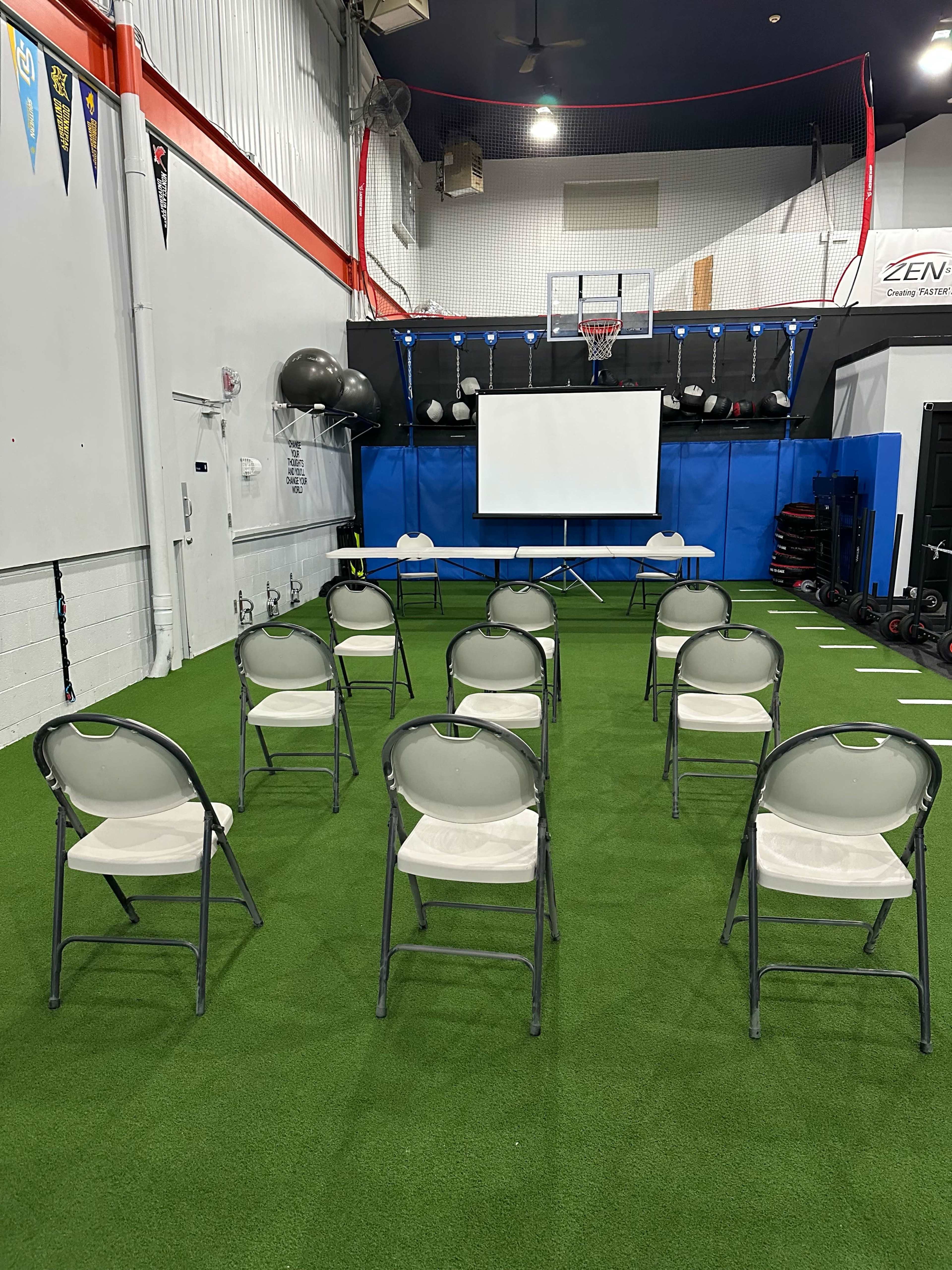 The image shows a gymnasium with a row of gray folding chairs arranged in front of a white screen and a basketball hoop in the background.
