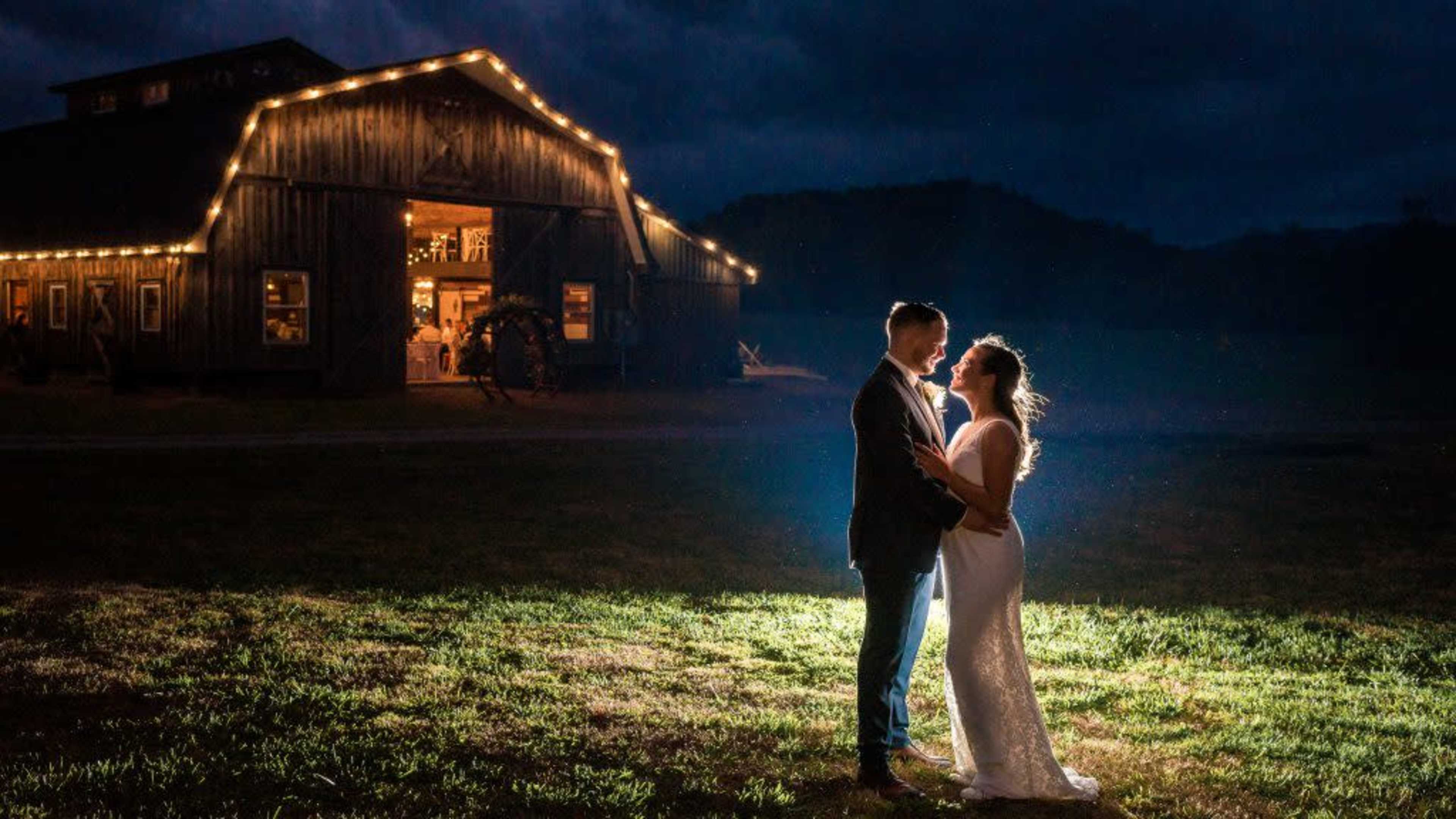 A couple stands close together under a spotlight in front of a barn illuminated with string lights.